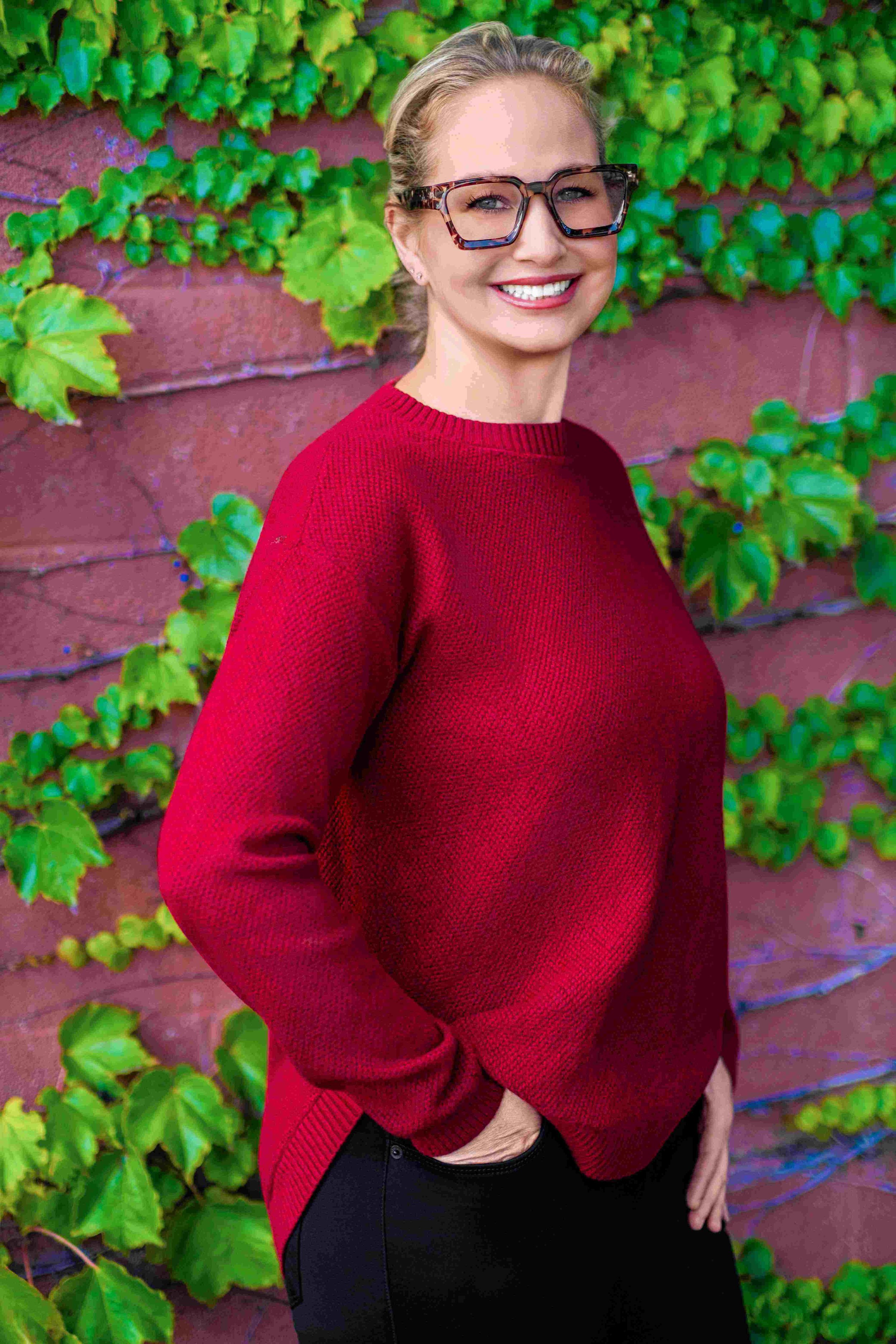A woman smiling and posing with her hand in her pocket in front of a brick wall with green vines and leaves. She is wearing her hair pulled up, a red sweater, and big square glasses.