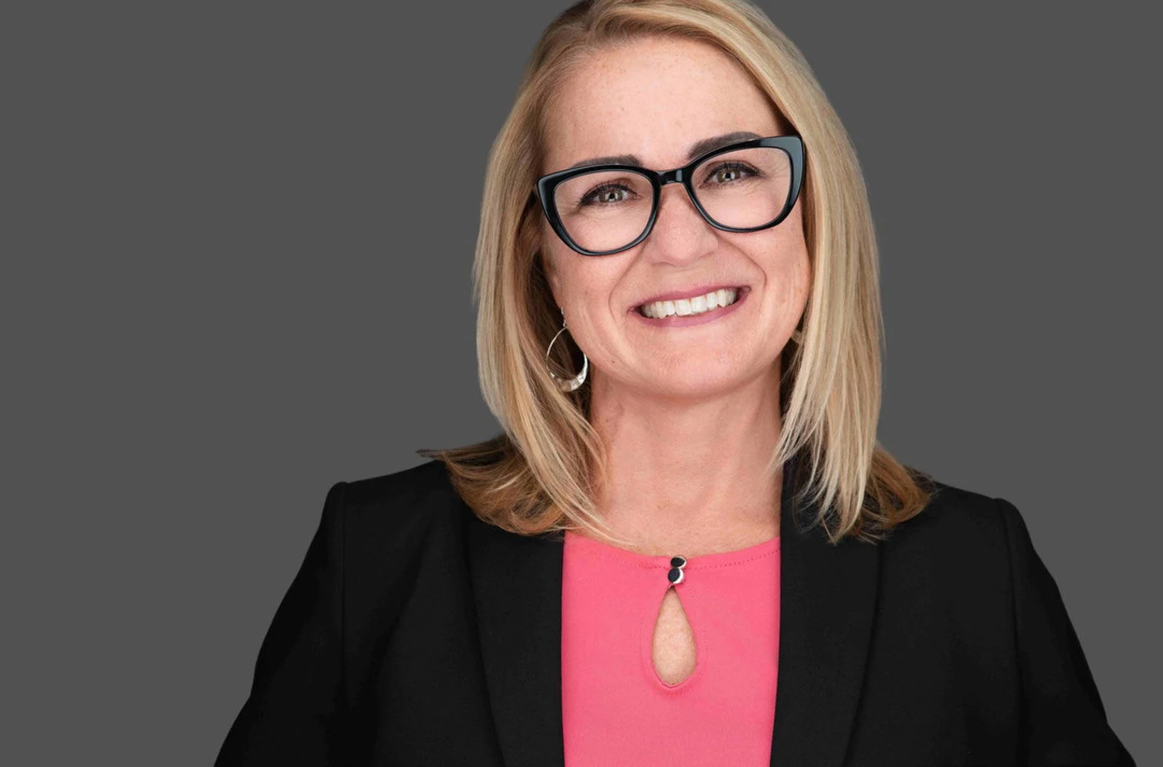 Headshot of a blonde woman wearing a black blazer, pink blouse, and black glasses.