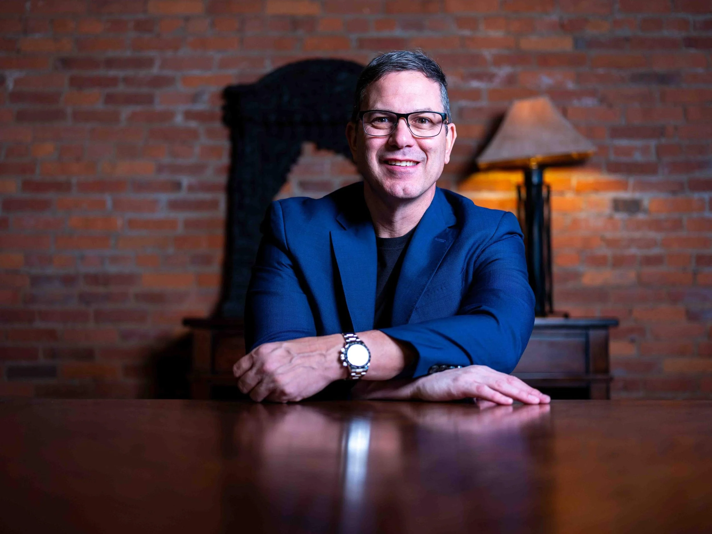Headshot of a man dressed in a blue suit with glasses and a watch. He is seated at a table in front of a dim lit brick wall