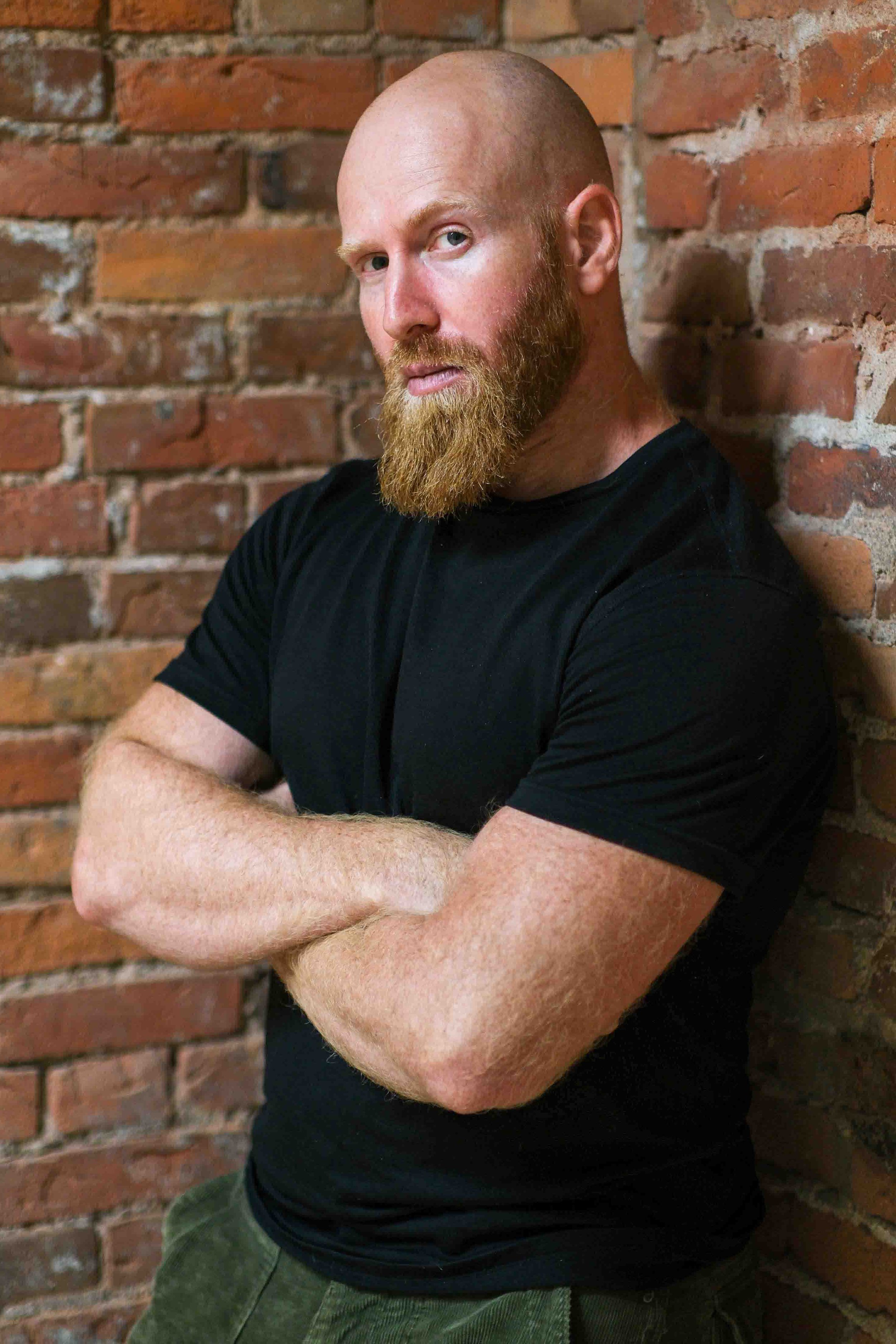 A bald man with a brown-orange beard posing with his arms crossed and wearing a black shirt with green pants.