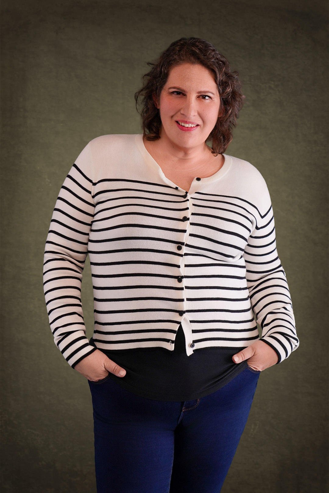 A woman with short brown hair posing for a headshot. She is wearing a white sweater with black stripes and skinny blue jeans, as well as placing her hands in her pockets and smiling.