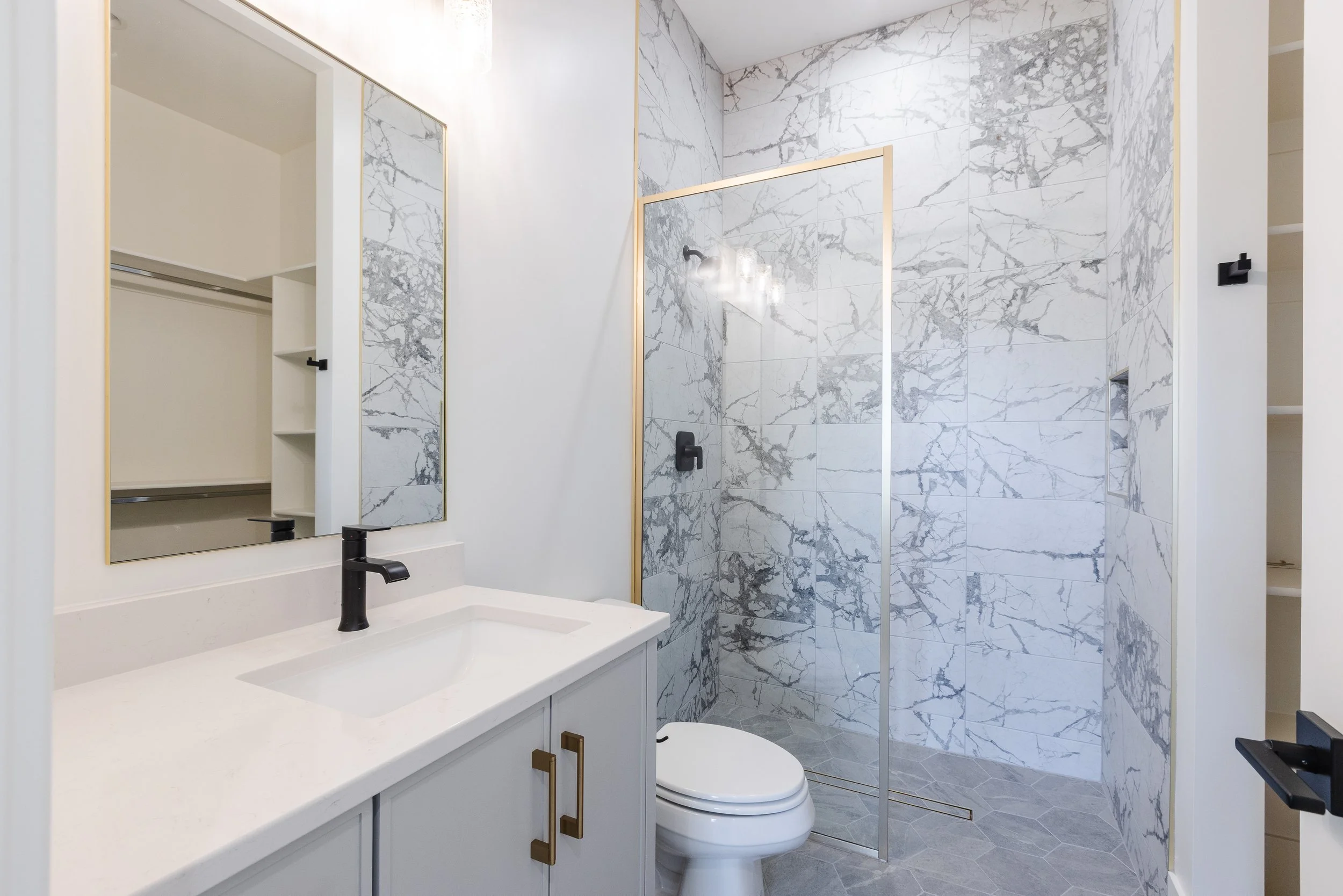 Modern bathroom with a marble shower, white vanity with black fixtures, mirror, and a toilet.