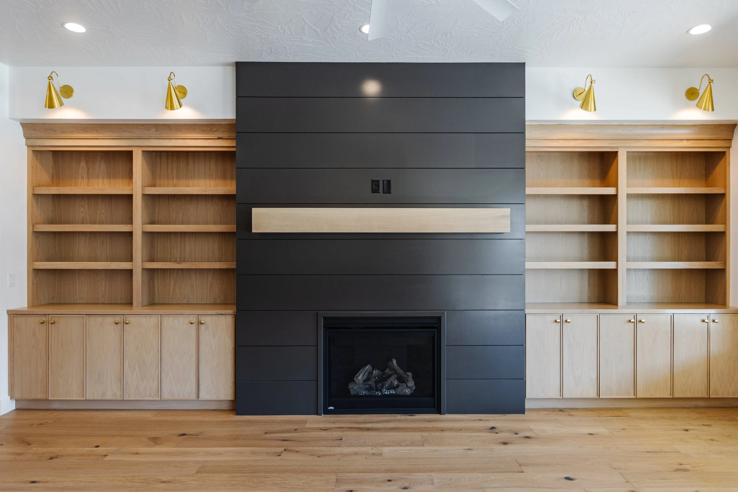 Empty built-in shelving units on either side of a modern black fireplace, with a large dark grey panel above the fireplace, and four gold art lamps installed on the white wall above the shelves.