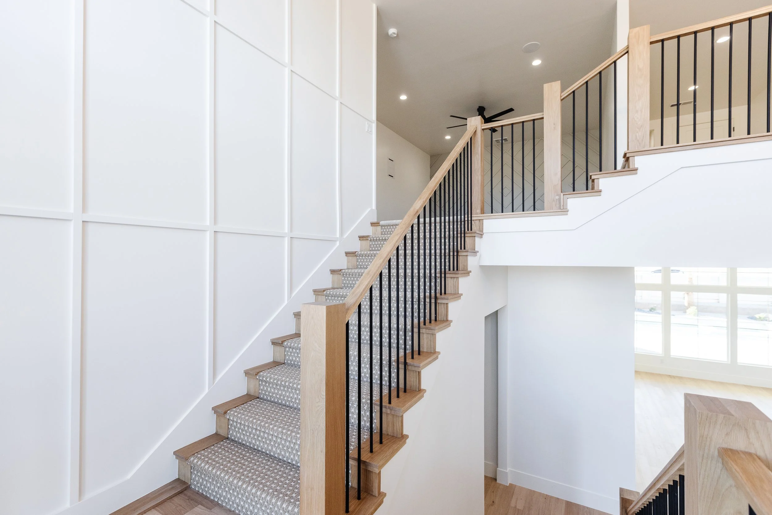 Interior of a modern house staircase with wood and black metal railing, patterned gray carpet on stairs, white walls, and windows letting in natural light.