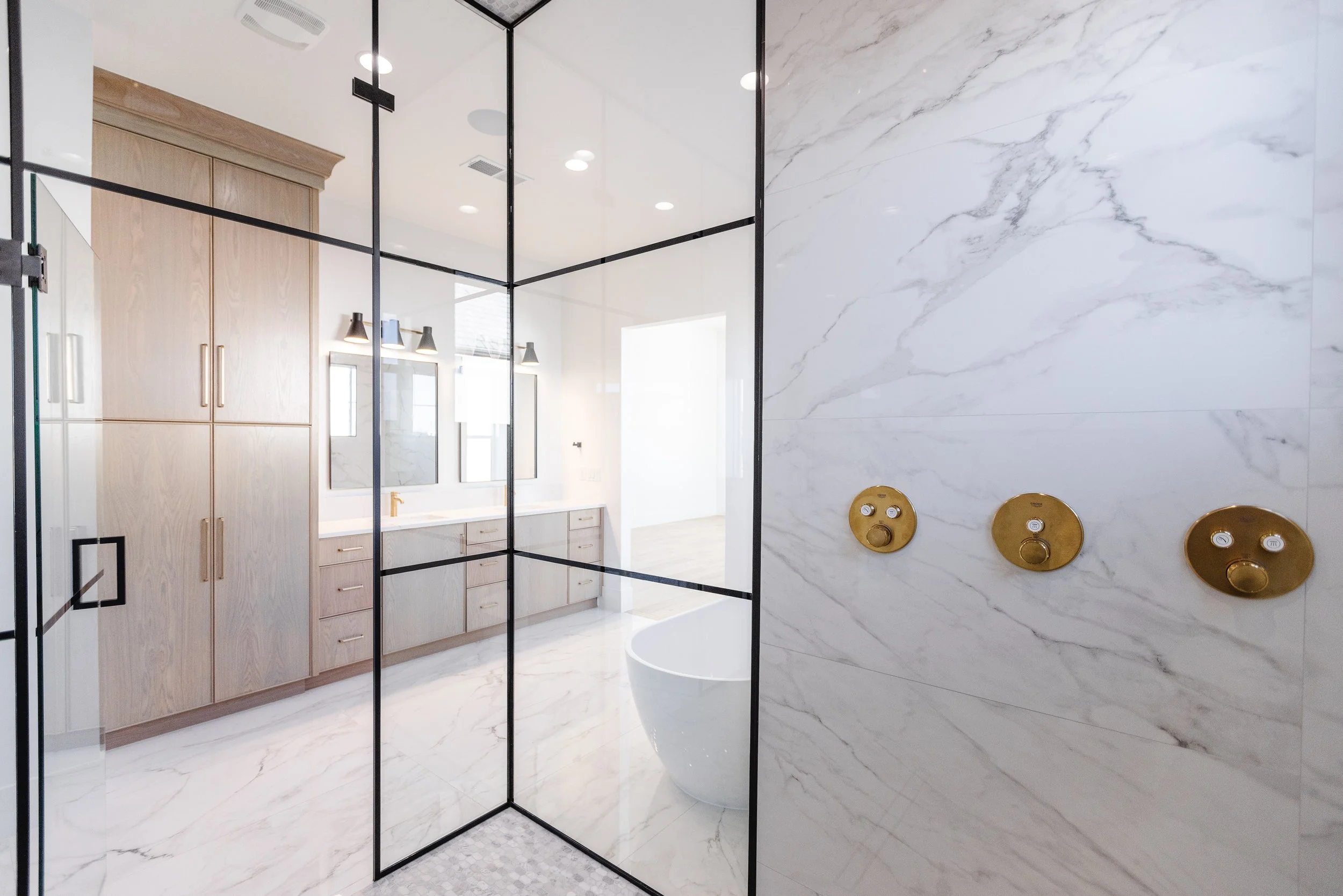 Modern bathroom with a glass shower door, marble wall, wooden cabinets, and gold fixtures.