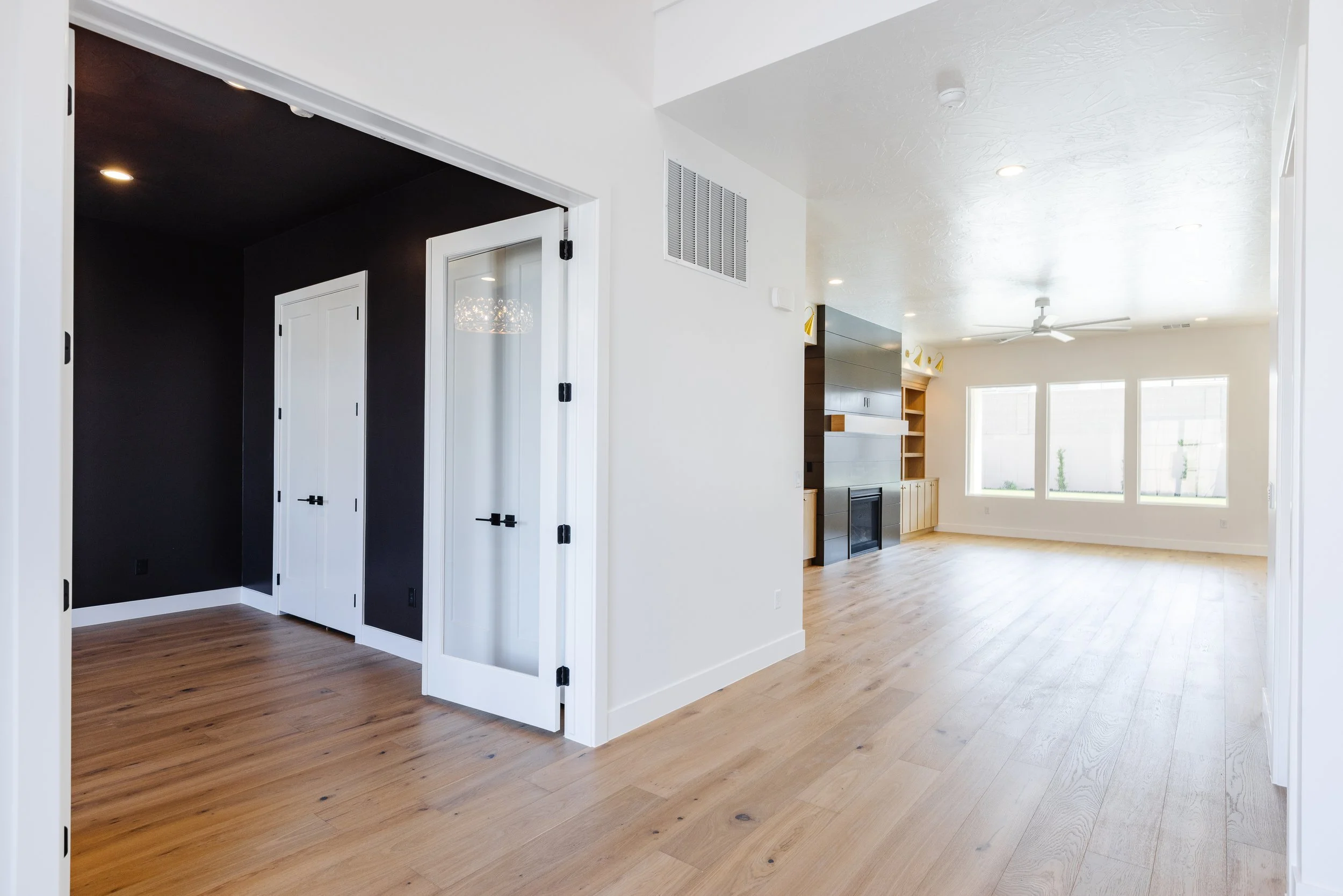 Bright living room with white walls, hardwood flooring, large windows, and a modern fireplace with dark wood paneling. An adjacent room with darker walls and framed doors is visible.
