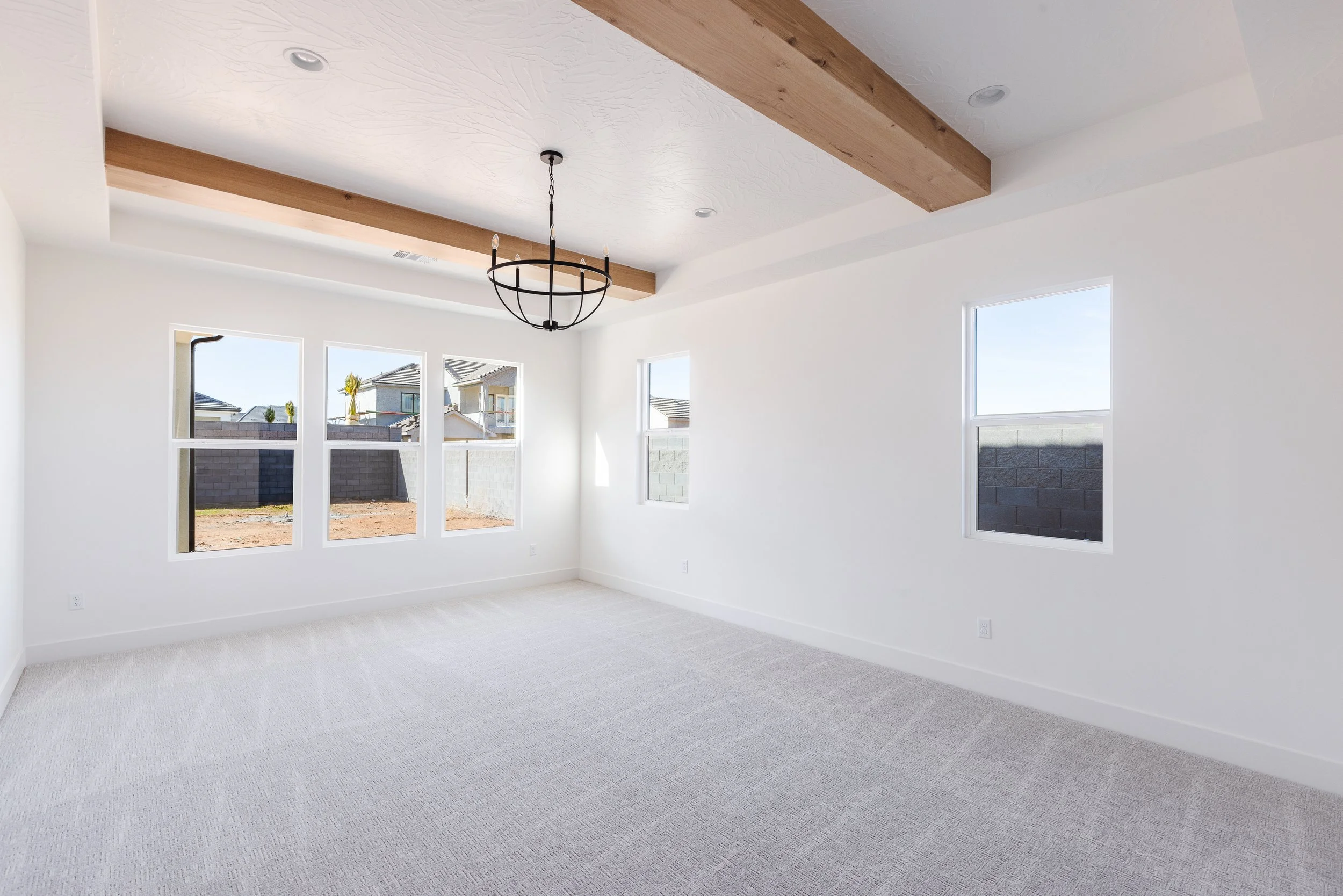 Empty living room with white walls, beige carpet, and multiple windows allowing natural light to enter. Wooden beams and a black chandelier are installed on the ceiling.