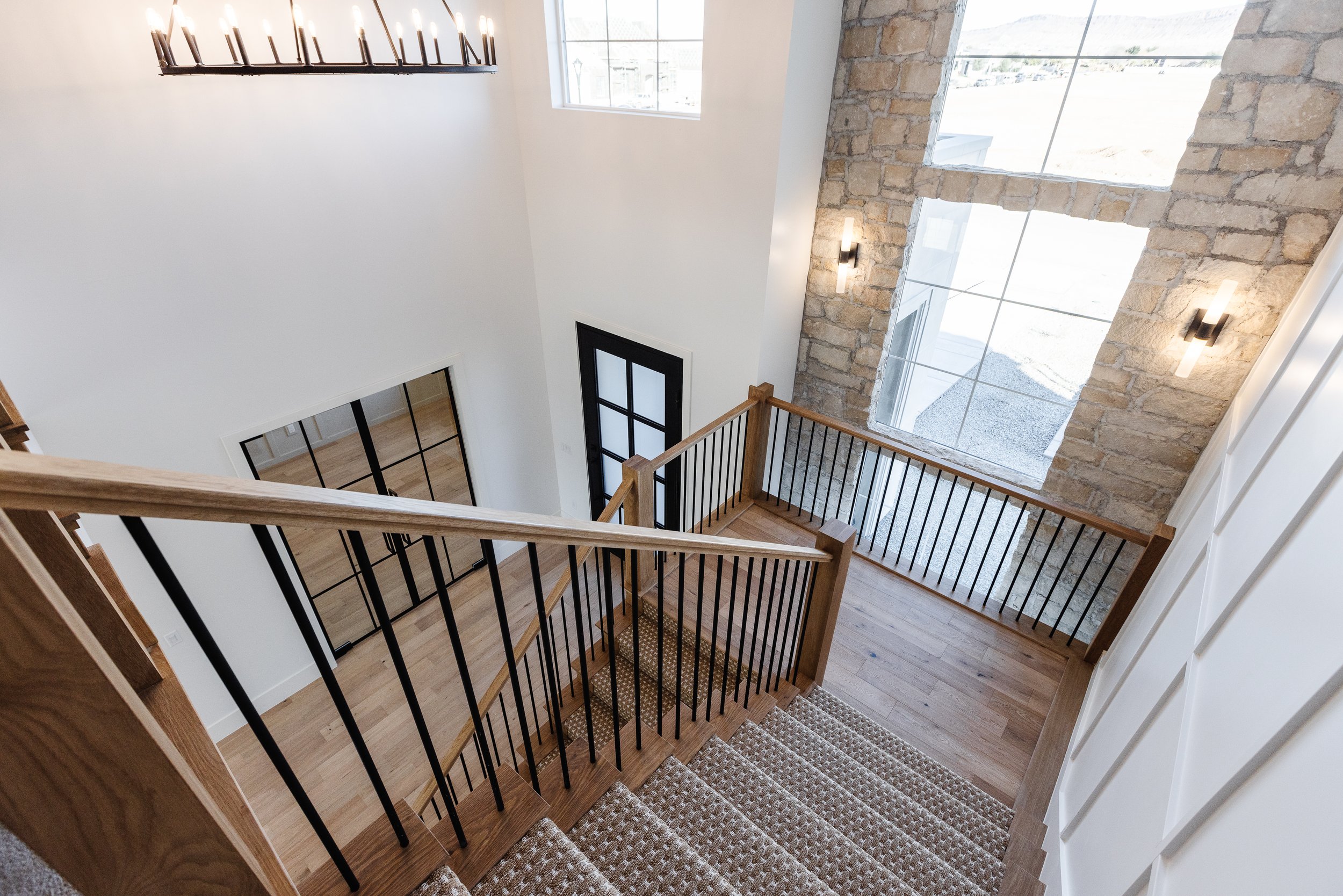 View of a staircase with wooden steps and black metal railings, leading to the second floor of a modern house featuring large stone and glass windows, with two wall-mounted light fixtures and a chandelier on the ceiling.