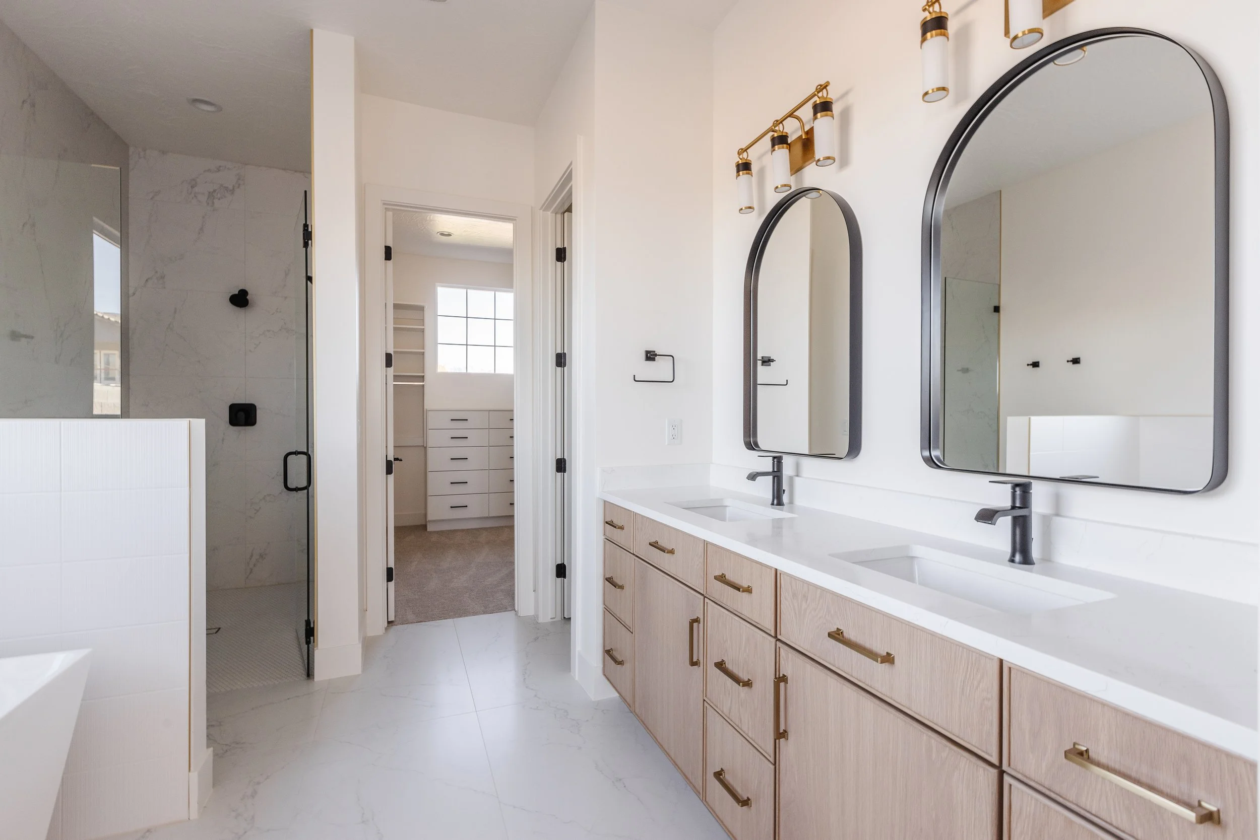 Modern bathroom with double sinks, light wood cabinets, black fixtures, large mirrors, and a walk-in shower with marble walls.