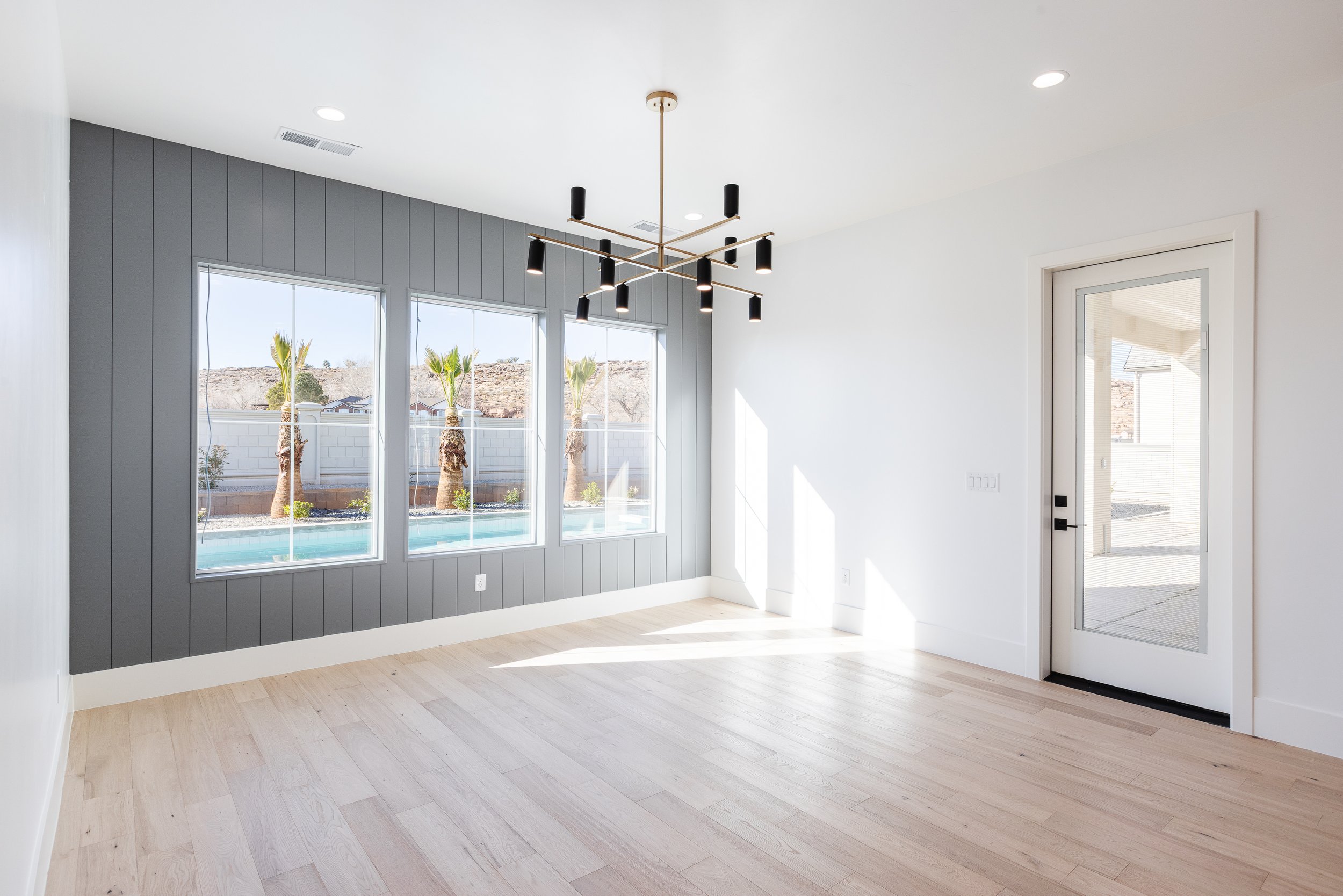 Empty room with large windows, modern black and gold chandelier, white walls, light wood flooring, and glass door leading outside.