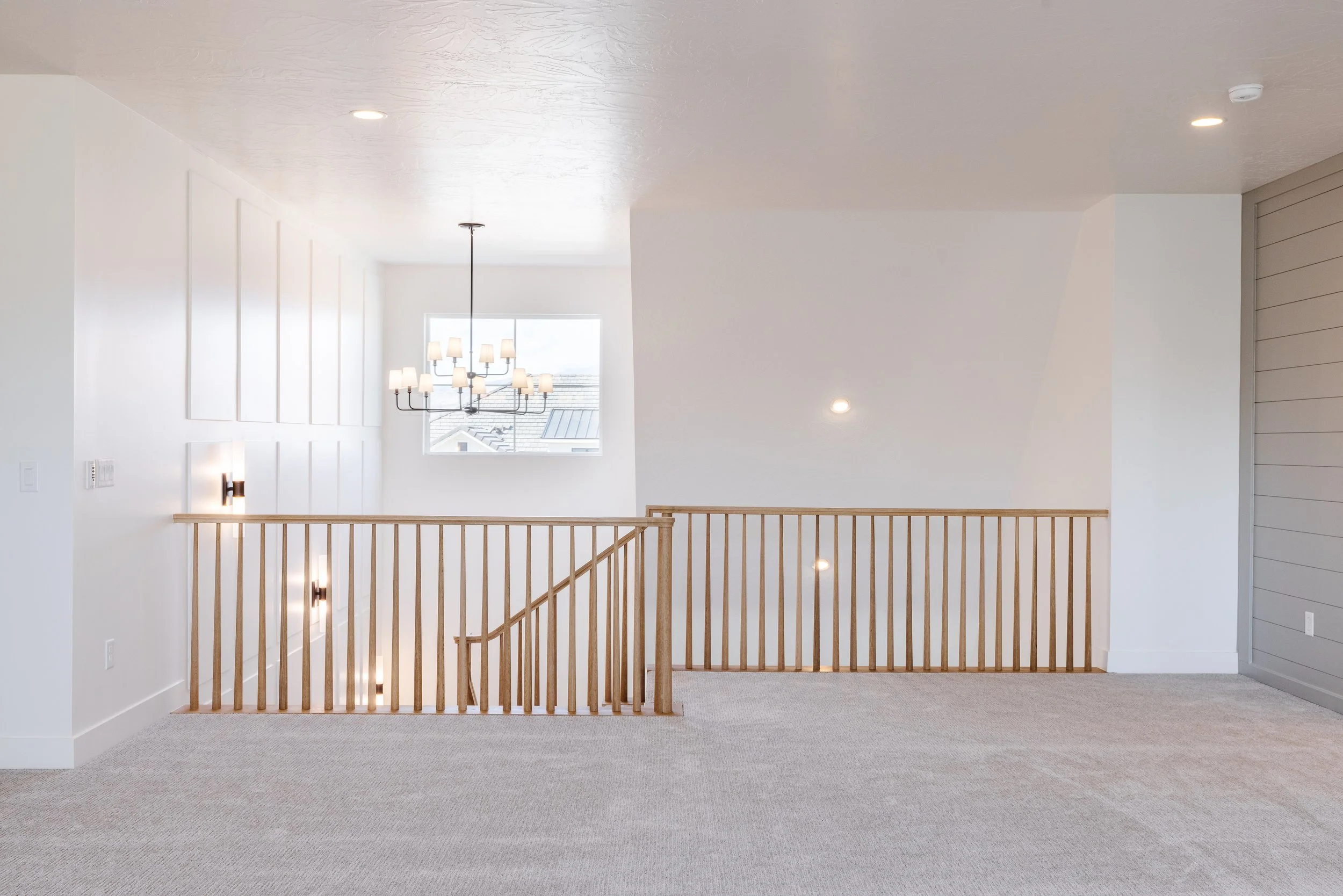 Interior view of a bright, modern second-floor space with white walls, carpeted floor, and a wooden railing overlooking the staircase below. A large window allows natural light to fill the area, and there is a chandelier hanging from the ceiling.
