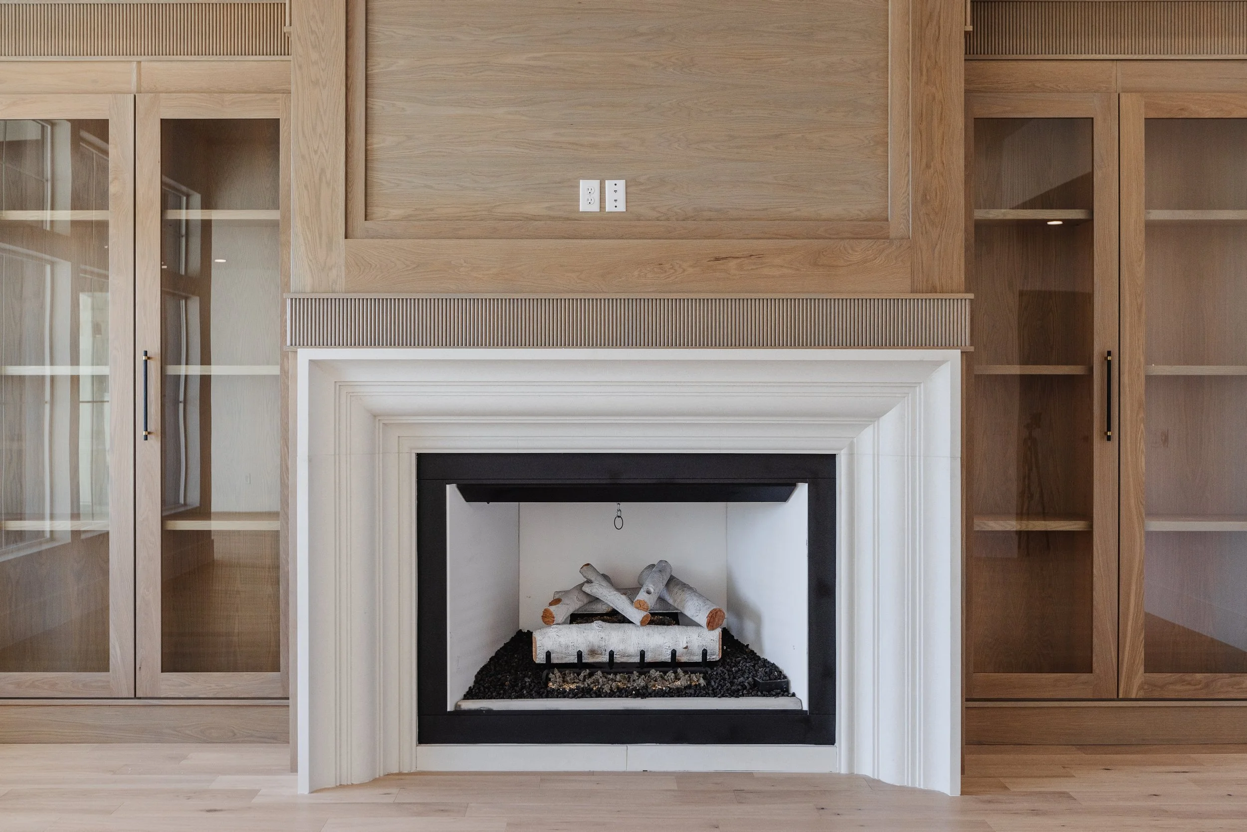 Empty fireplace with a white mantel and black trim, surrounded by built-in wooden cabinets with glass doors on both sides, in a wood-paneled room.