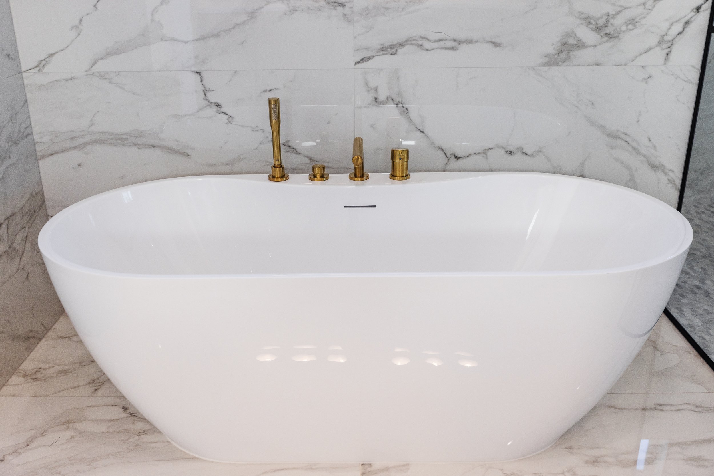 White freestanding bathtub with gold fixtures in a bathroom with marble walls and floor.
