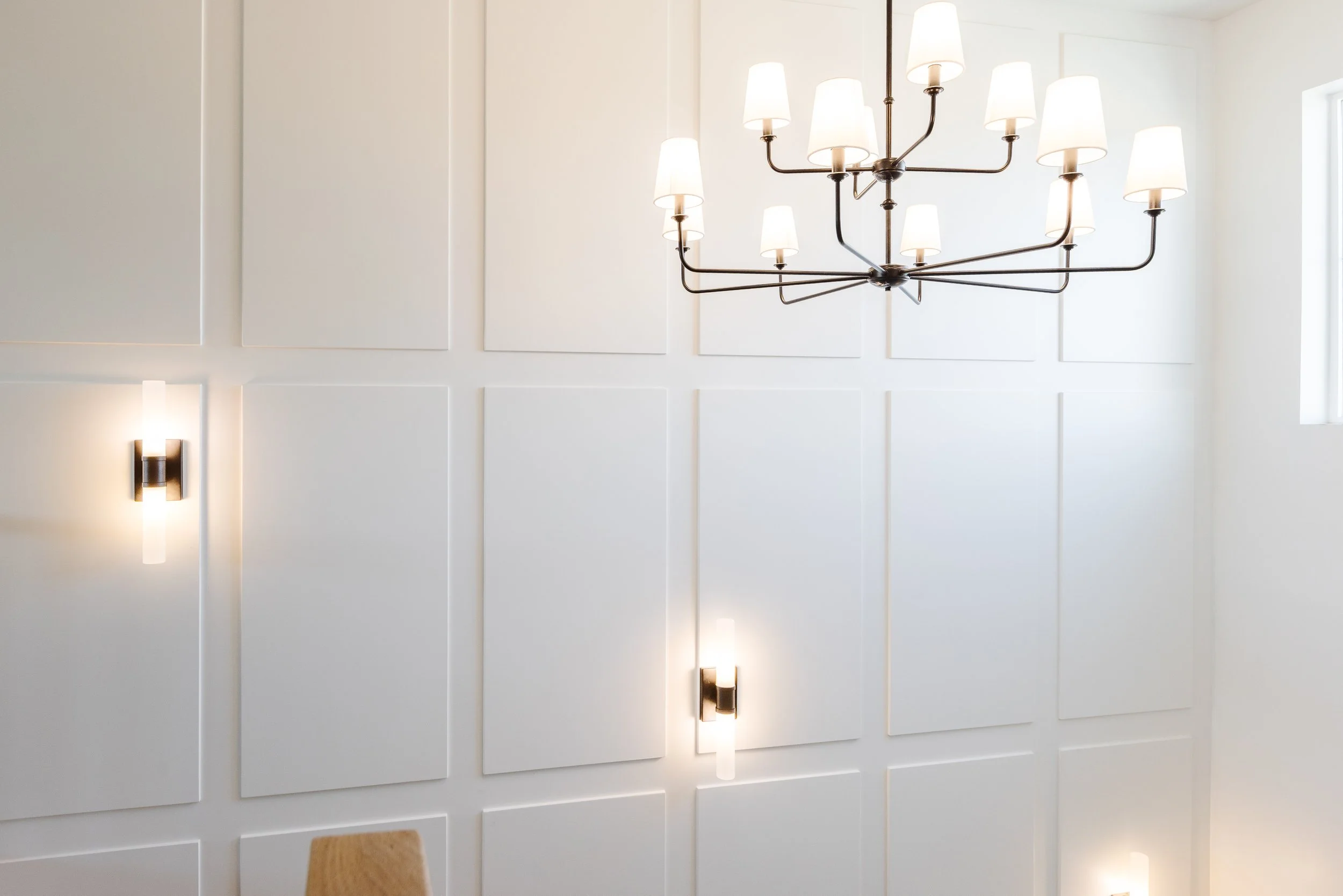 Modern chandelier with multiple white lampshades hanging from a metallic frame in a bright room with white paneled walls and wall sconces.