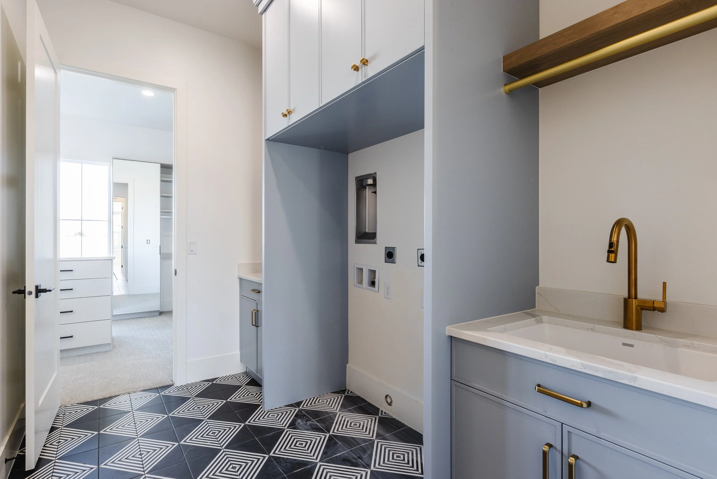 Modern kitchen with blue and white cabinetry, black patterned floor tiles, gold hardware, and a white countertop with a gold faucet. An open doorway leads to a bright room with white walls and built-in shelves.