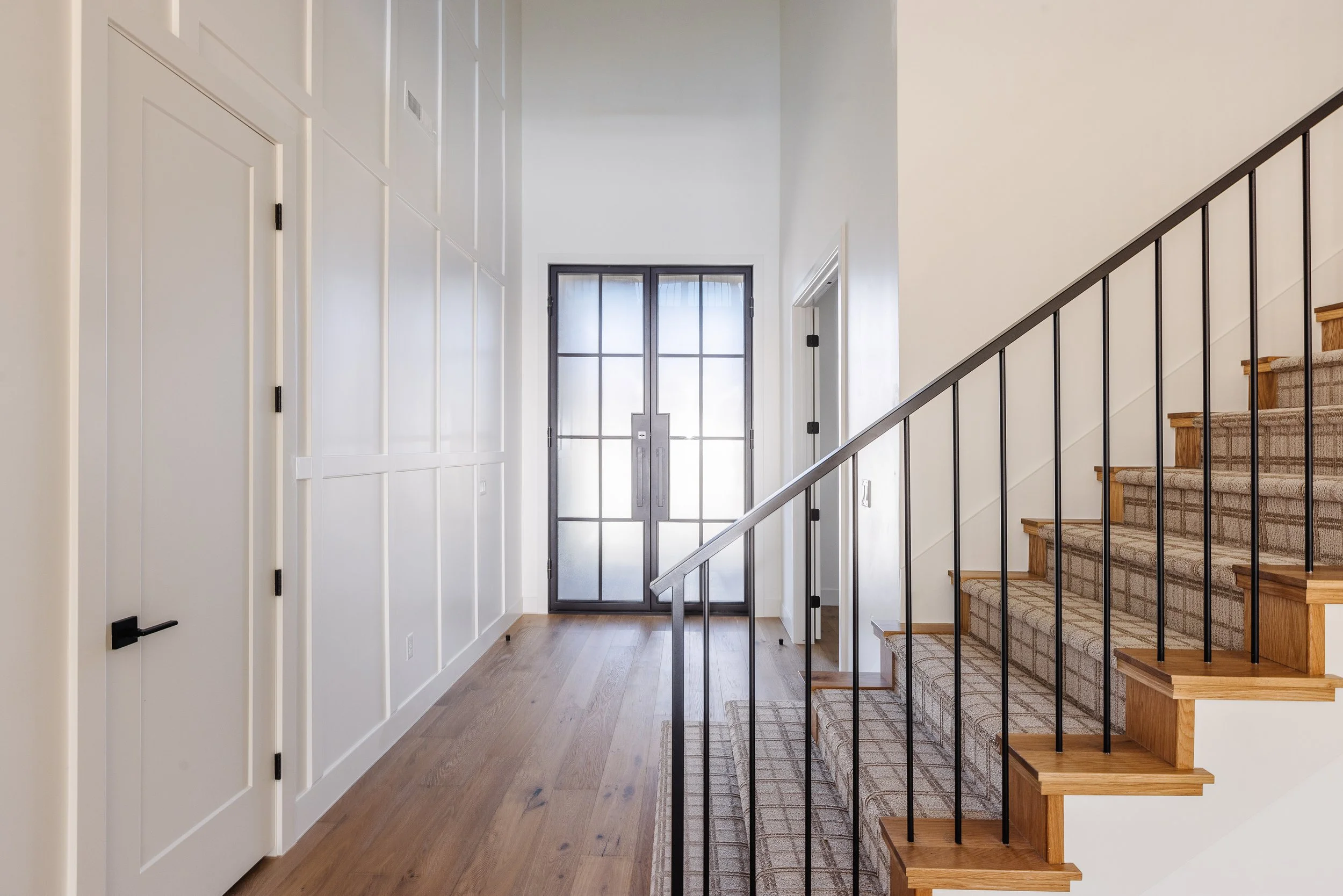 Entryway with a glass-paneled front door, wooden stairs with black metal railing, white paneled closet, and hardwood floors.