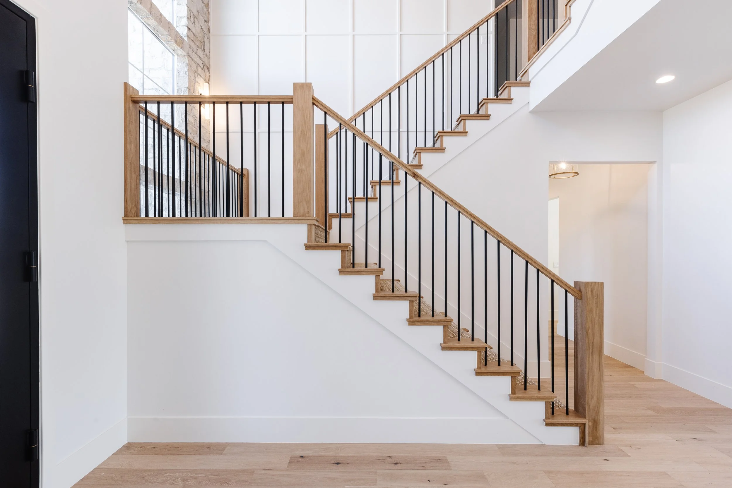 Interior of a modern house with a wooden staircase, black metal railing, white walls, a stone accent wall near a window, hardwood floors, and minimalistic lighting.