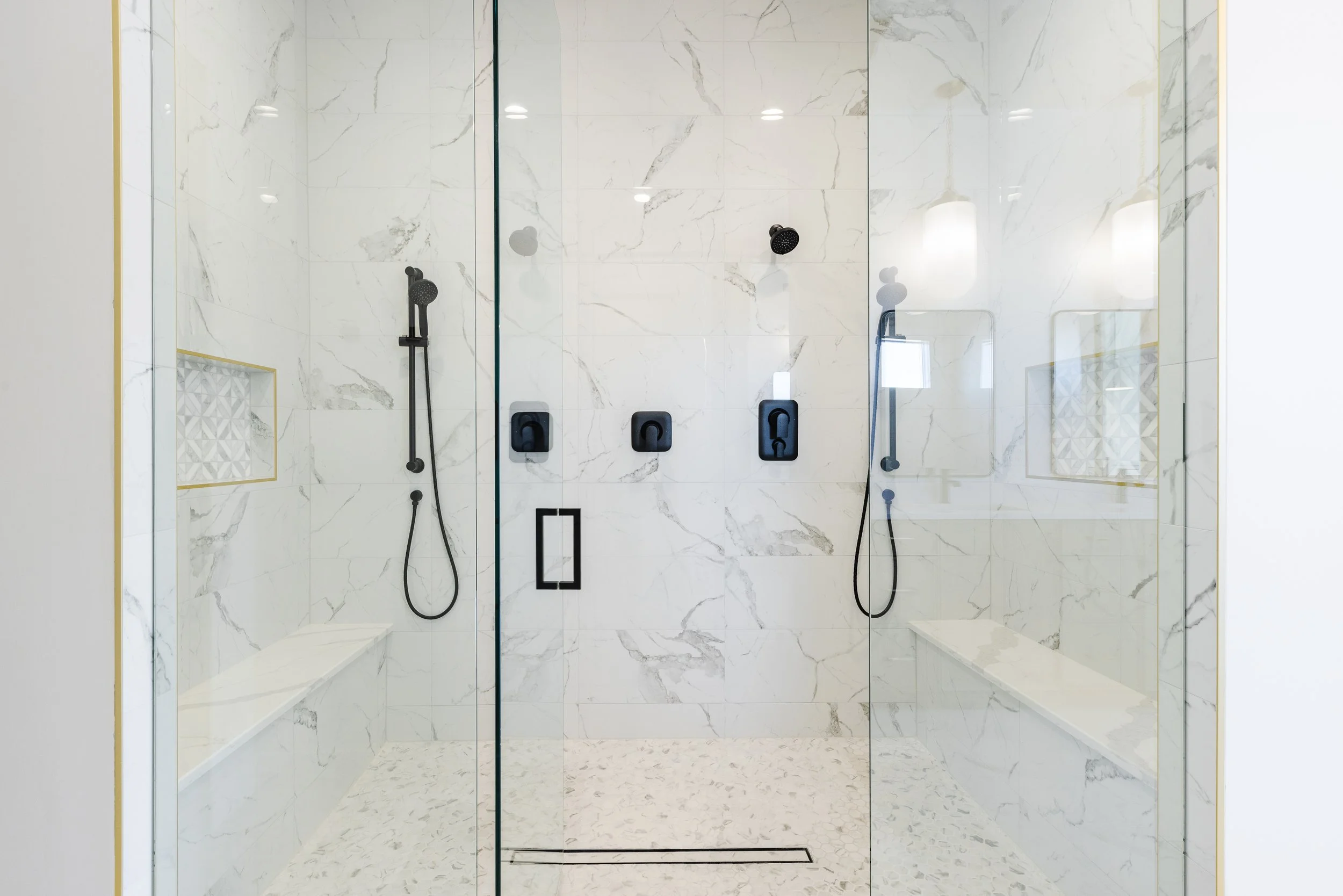 A modern glass-enclosed shower with marble wall and floor tiles, two handheld showerheads, and built-in benches.