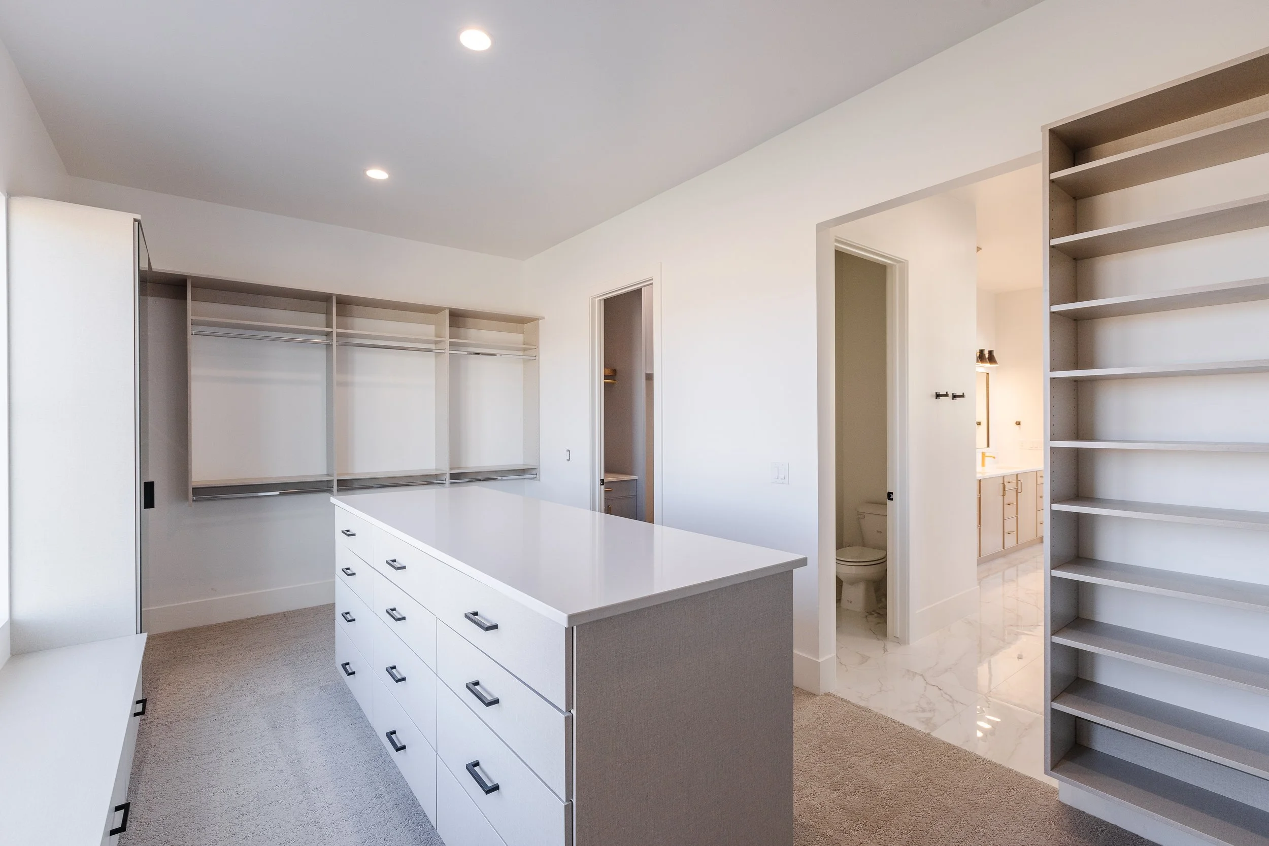 Empty walk-in closet with white drawers, open shelves, and a nearby bathroom with marble flooring and a visible toilet.