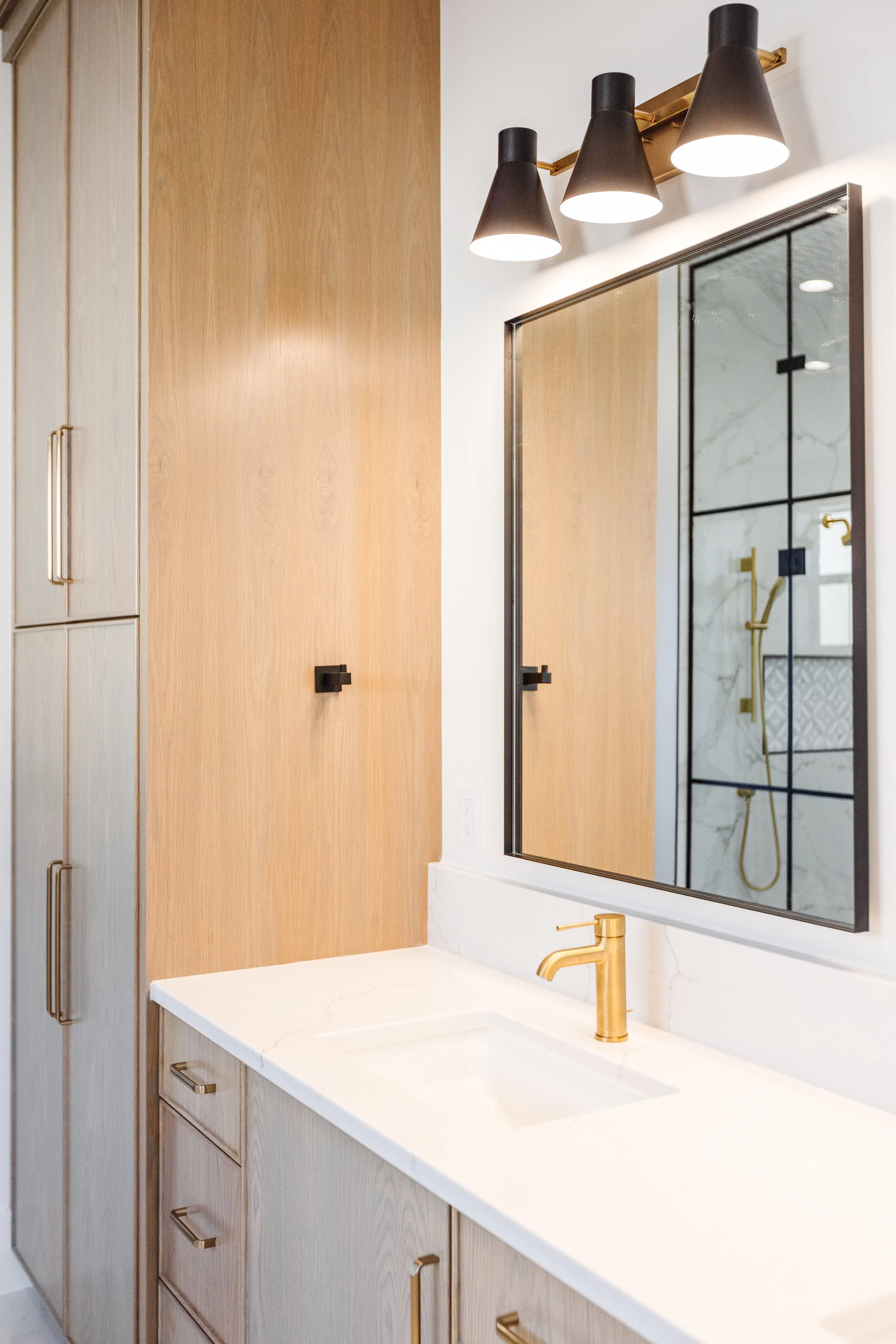 A modern bathroom with light wood cabinets, a white marble countertop, a large mirror, and black and gold fixtures, including a gold faucet and black handles. A black wall-mounted light fixture with three cone-shaped shades is visible, and a shower w