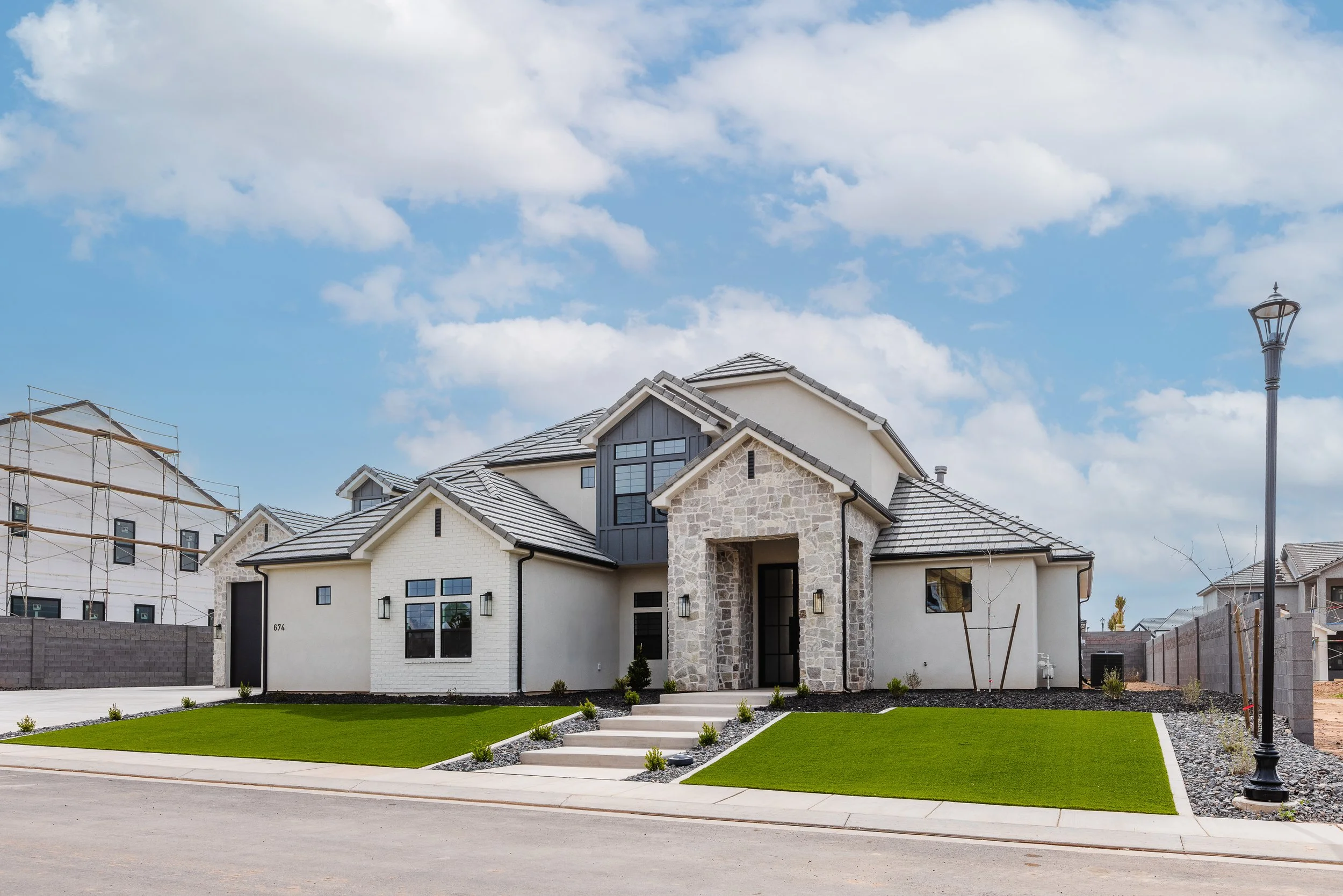 Modern two-story house under construction with gray tiled roof, stone and white exterior walls, front yard with green grass, concrete steps, and black fence. Construction scaffolding visible on neighboring building. Clear blue sky with clouds.