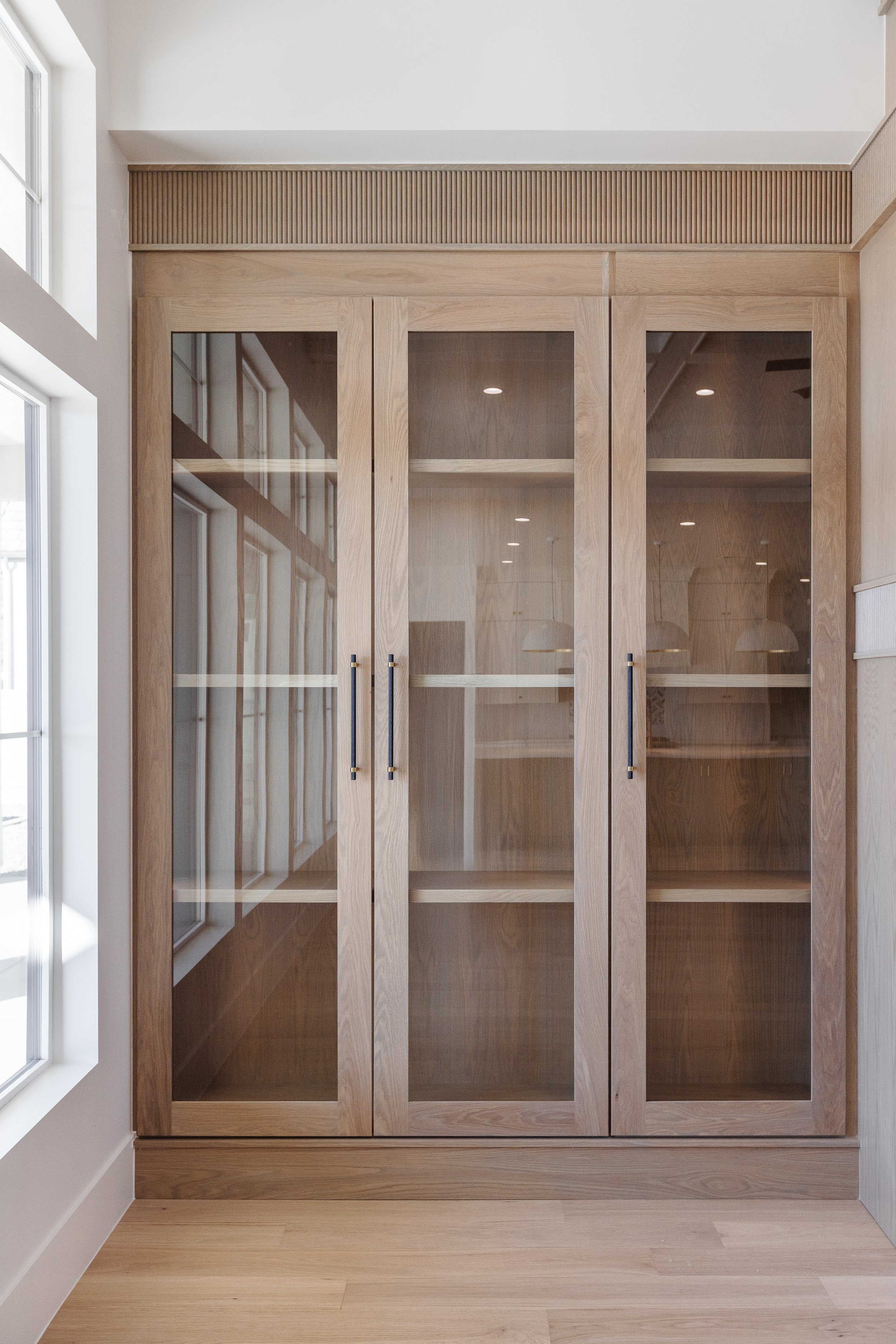 Interior view of a large wooden cabinet with glass doors, located next to a window, with a light wood floor and ceiling details.