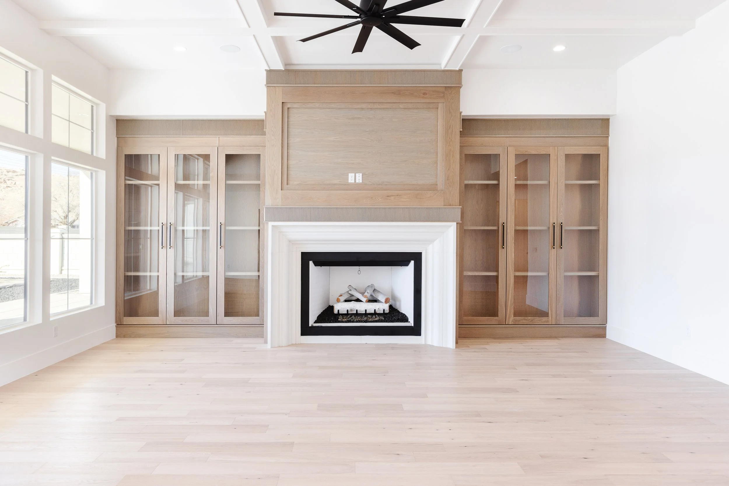 Living room with a fireplace, built-in wooden bookcases, large windows on the left, and a black ceiling fan.