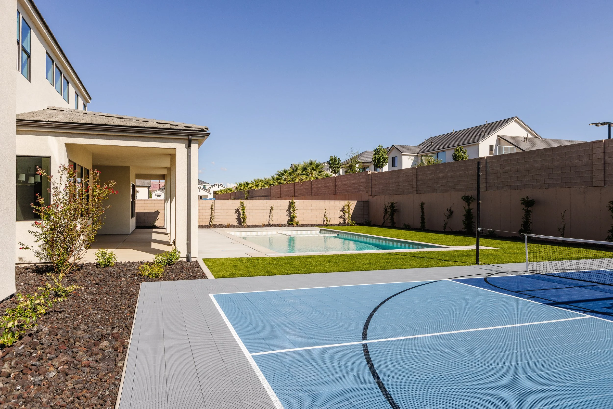 A backyard with a swimming pool, a sports court, and surrounding houses with trees and a brick wall.