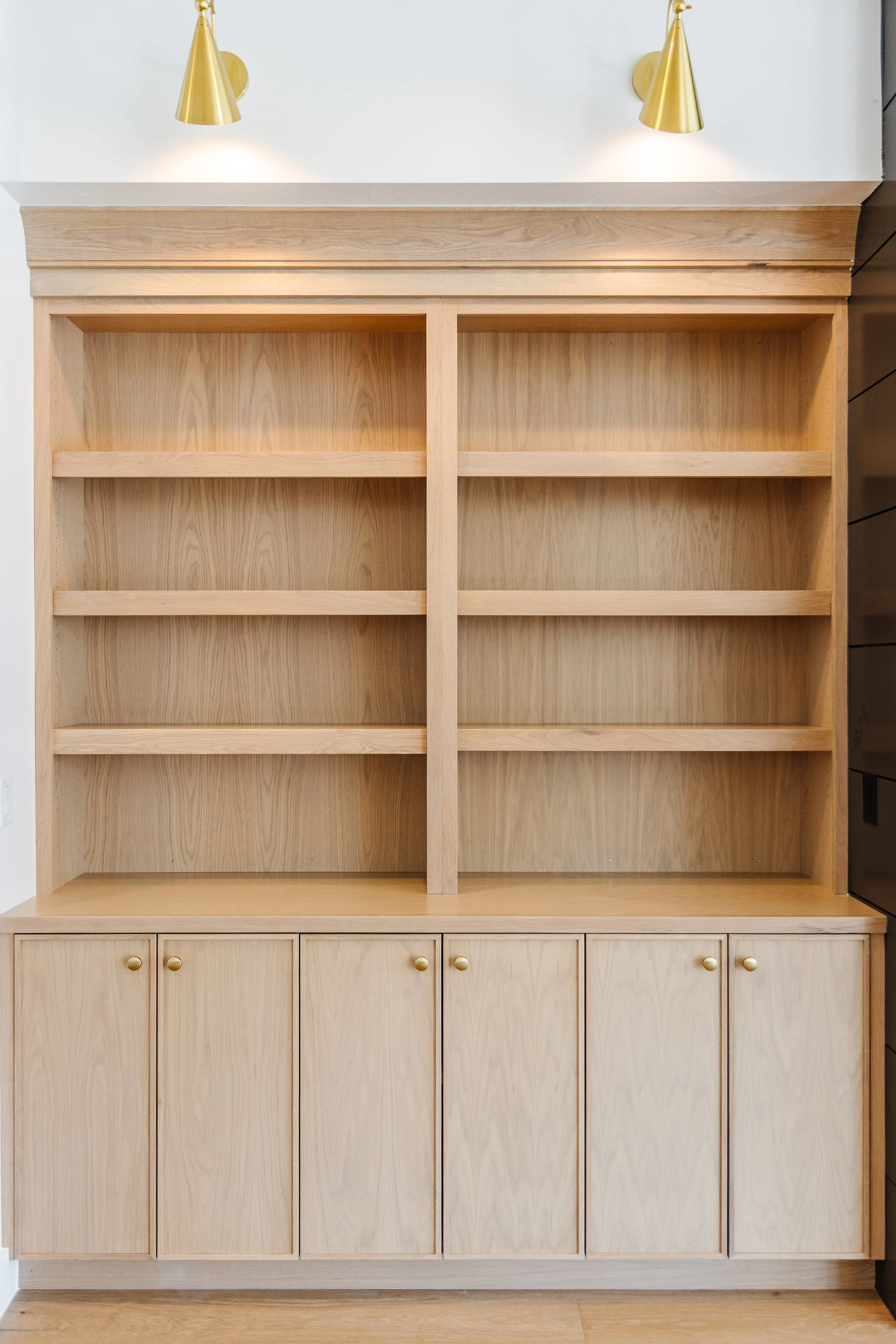 Empty light wood bookshelf with cabinet drawers at the bottom, top part has four open shelves, mounted on white wall with modern gold wall sconces above.