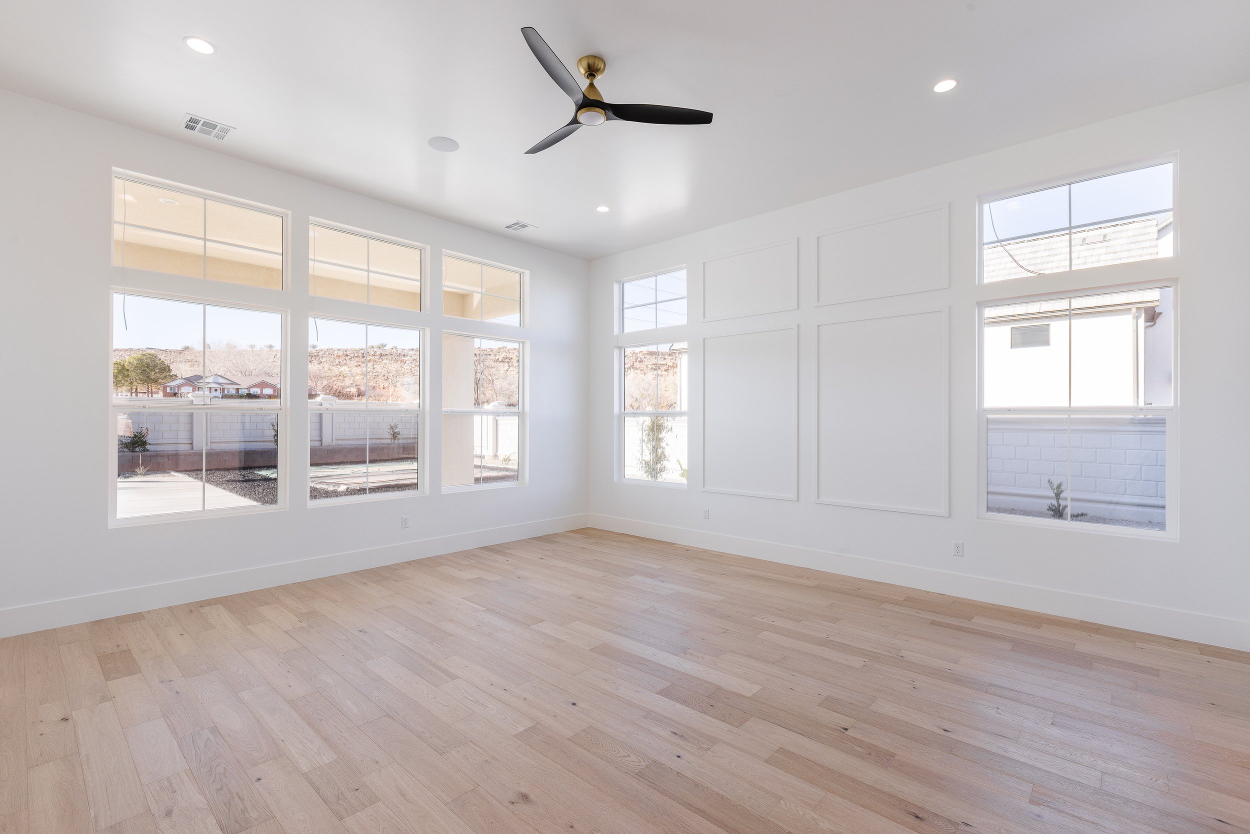Empty room with large windows, light-colored hardwood floors, white walls, ceiling fan, and recessed lighting.
