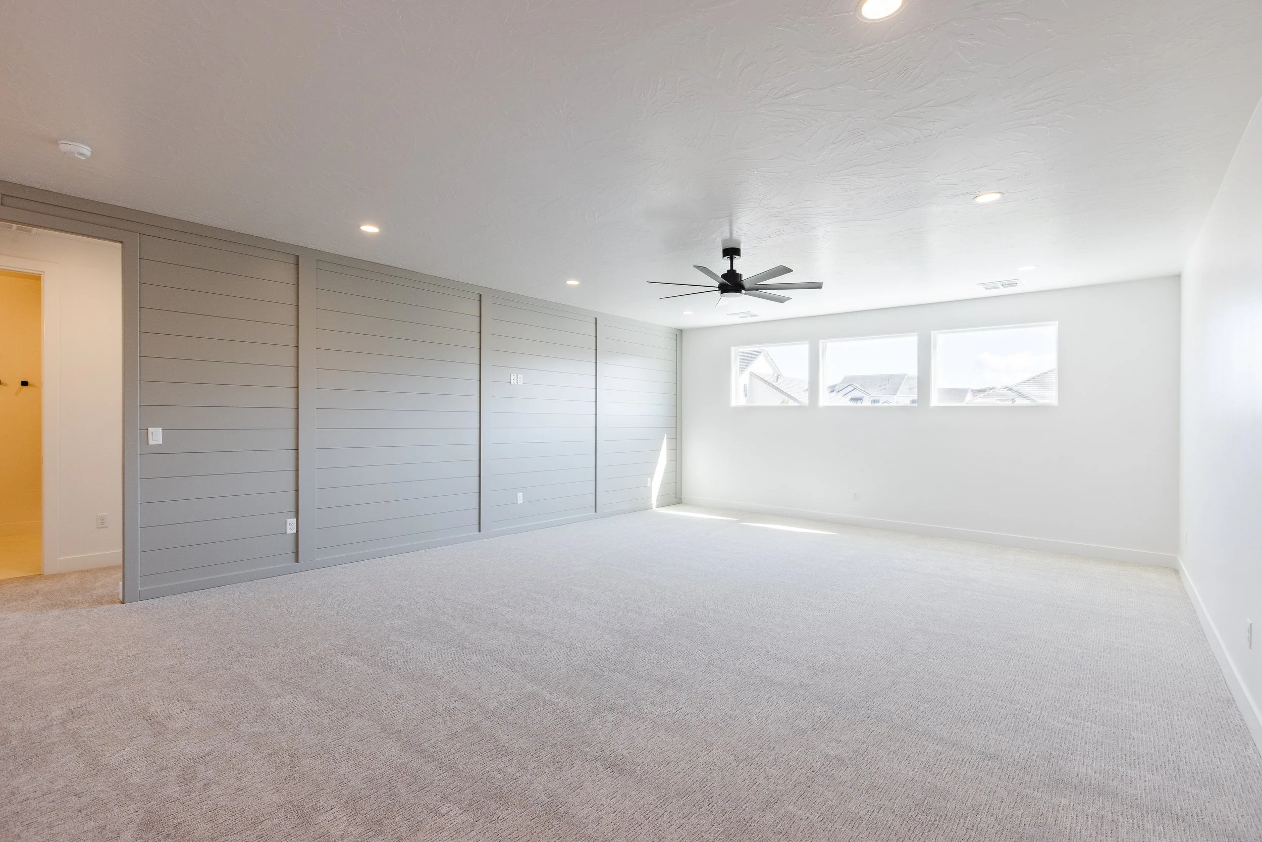Empty room with beige carpet, gray shiplap wall, white walls, ceiling fan, and three windows letting in natural light.