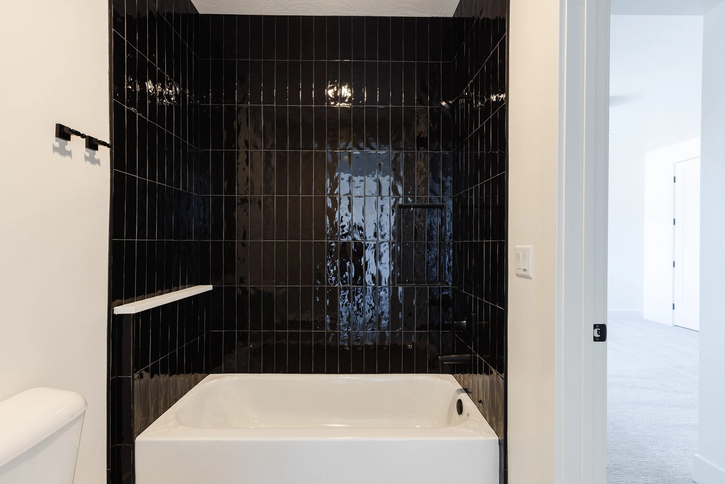 Empty black tiled shower with a built-in shelf, a showerhead, and a white bathtub base, next to a white wall and doorway.