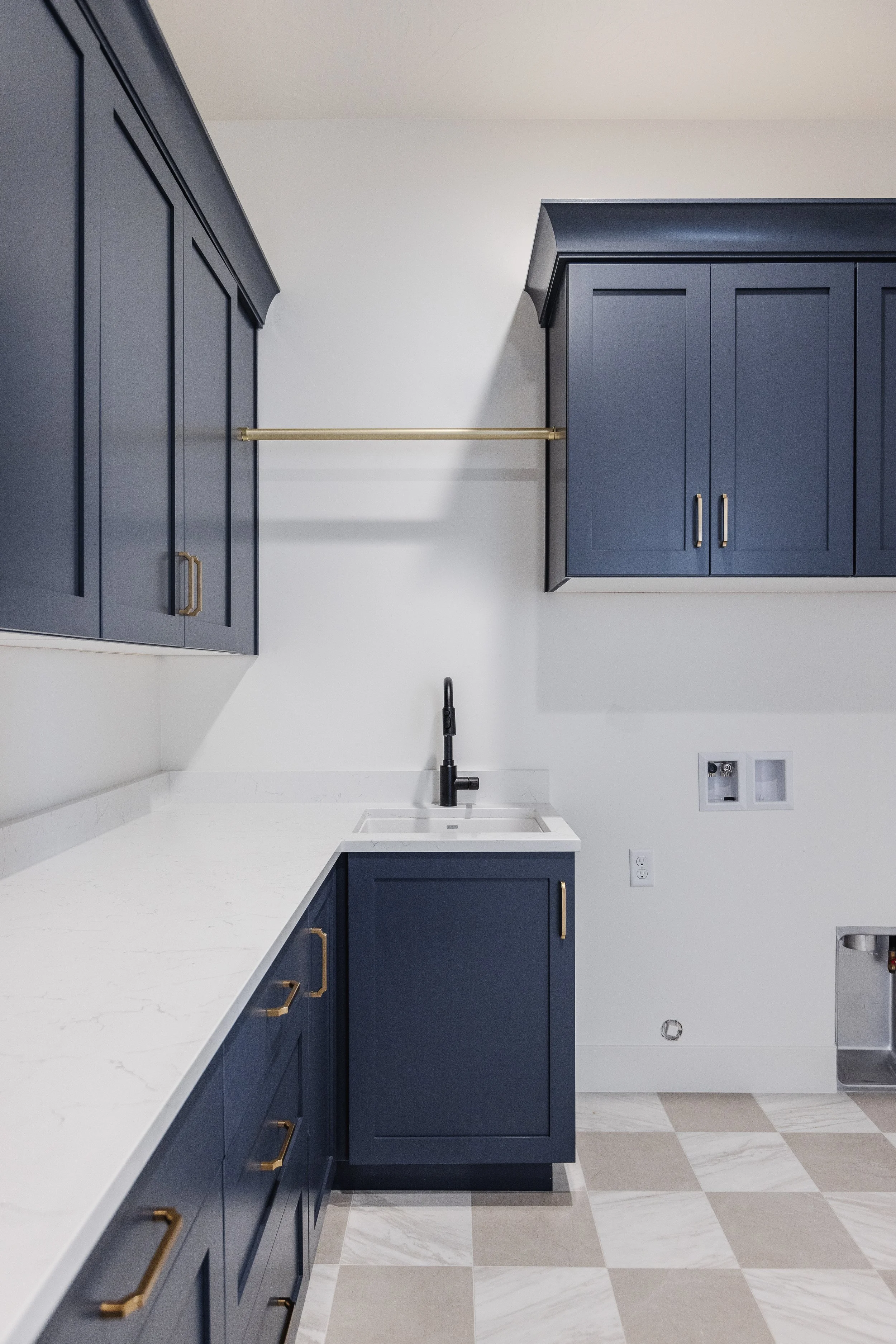 Modern kitchen with navy blue cabinets, gold handles, white marble countertop, black faucet, and white walls. There's a gold bar for hanging items on the upper wall and a laundry or utility area with outlets and a washing machine vent.