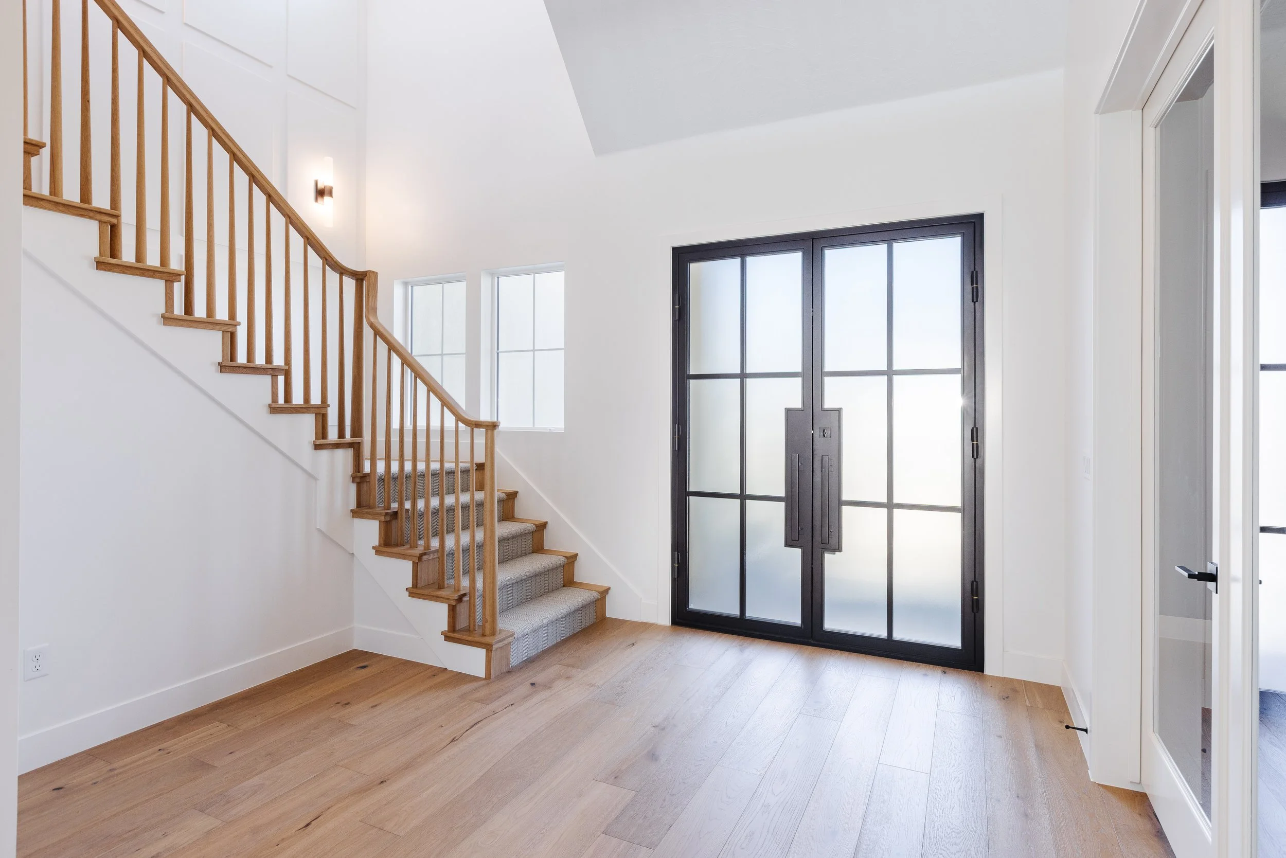 Entryway with a staircase on the left, a large black front door with glass panes, and white walls and trim. Wooden flooring and a window near the stairs.