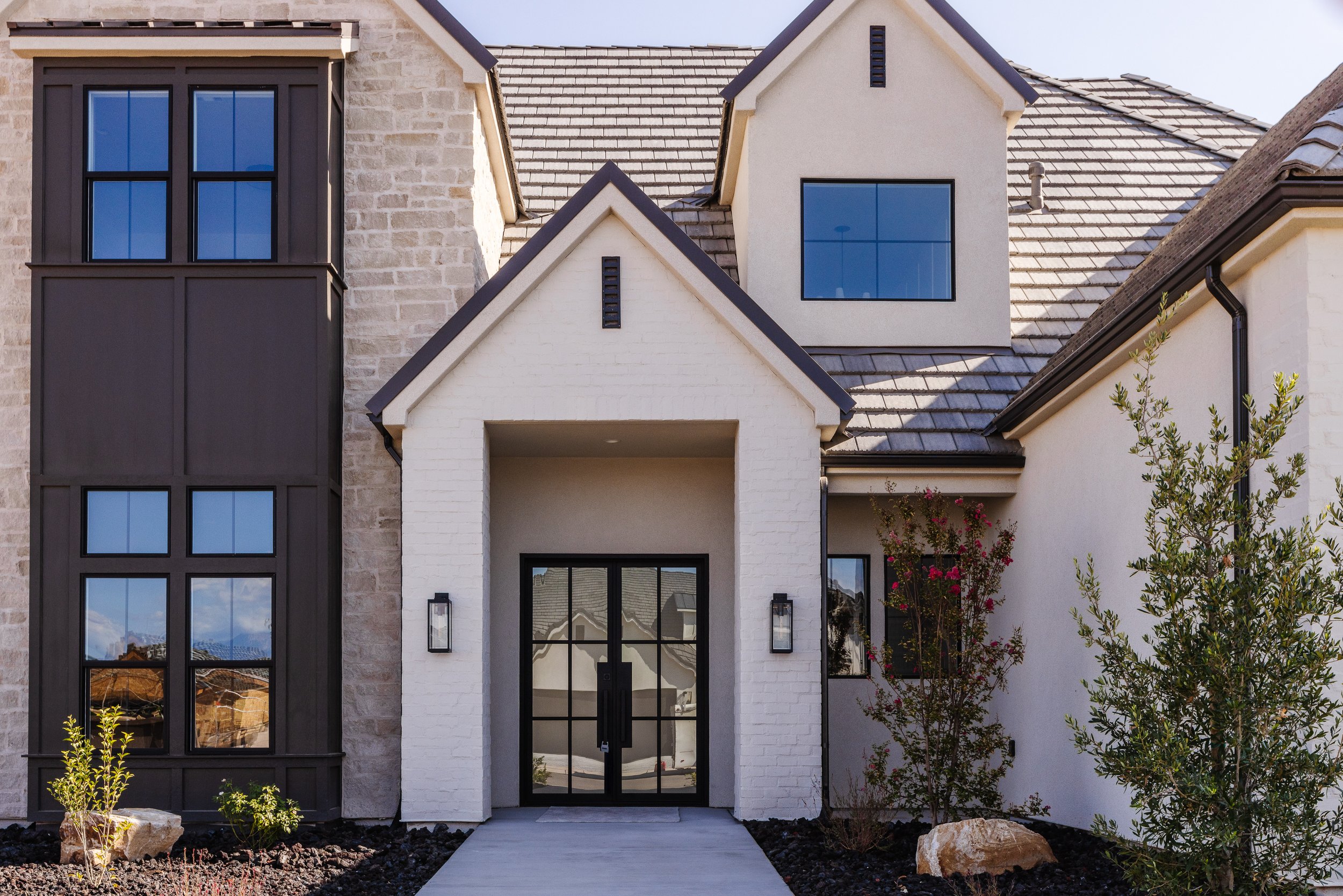 Front view of a modern house with a combination of brick, white stucco, and dark exterior panels, large windows, and a landscaped front yard with rocks and plants.