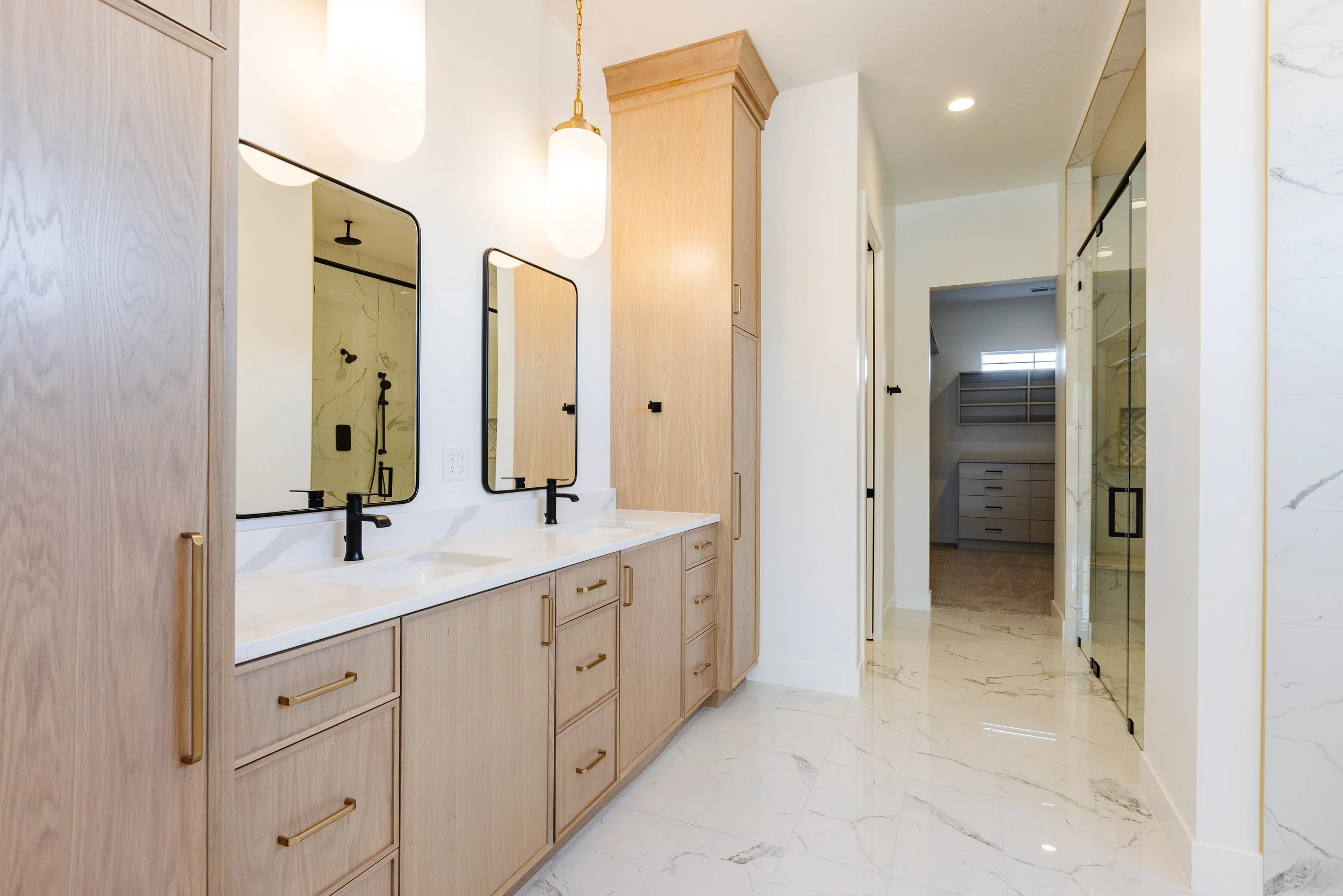 Modern bathroom with dual sinks, black faucets, wooden cabinetry with gold handles, large marble tile floors, walk-in shower with glass door, and reflective mirrors.