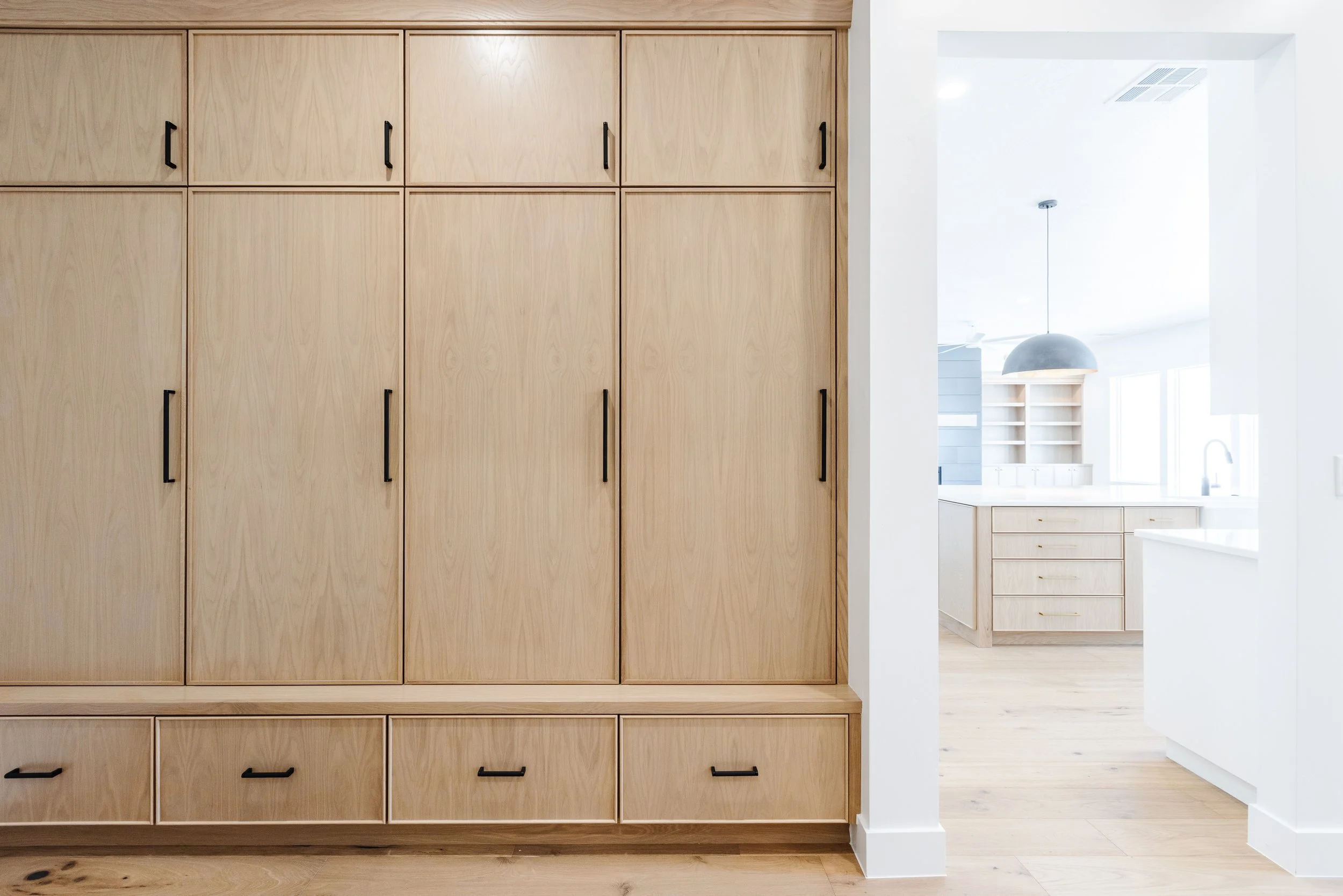 Kitchen with large wooden cabinets and black handles, white walls, light wood flooring, and a partial view of a kitchen island with drawers and a pendant light in the background.