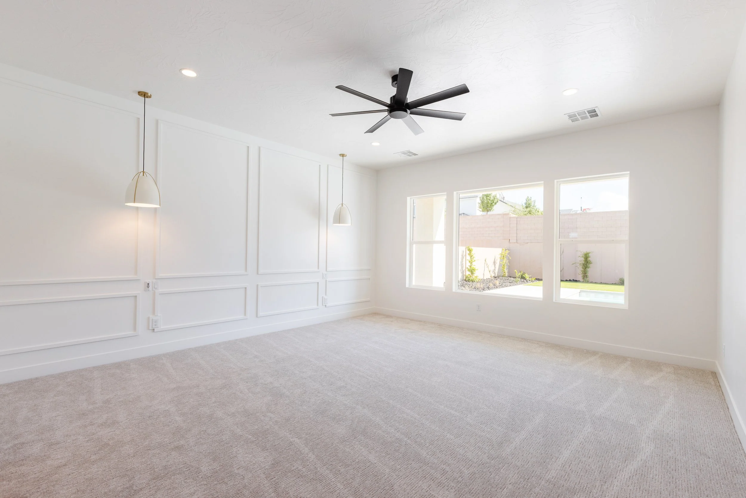 Empty living room with large windows, white walls, beige carpet, ceiling fan, and pendant lights.