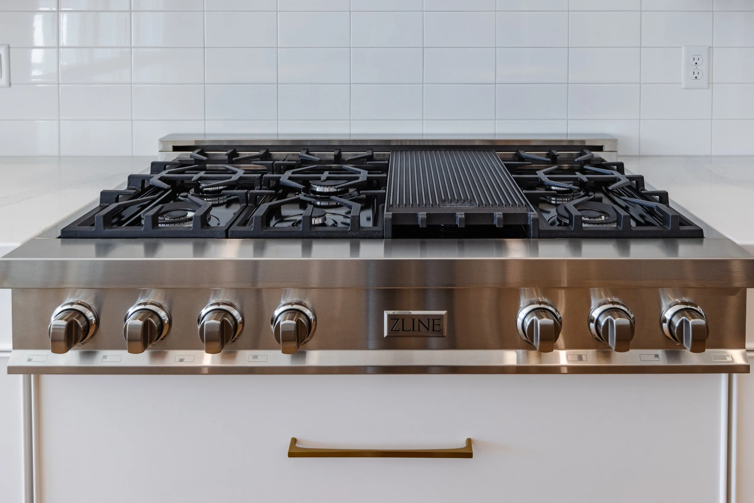 Stainless steel ZLINE gas range with six burners, a griddle in the middle, and control knobs at the front, set against a white tiled wall with electrical outlets.