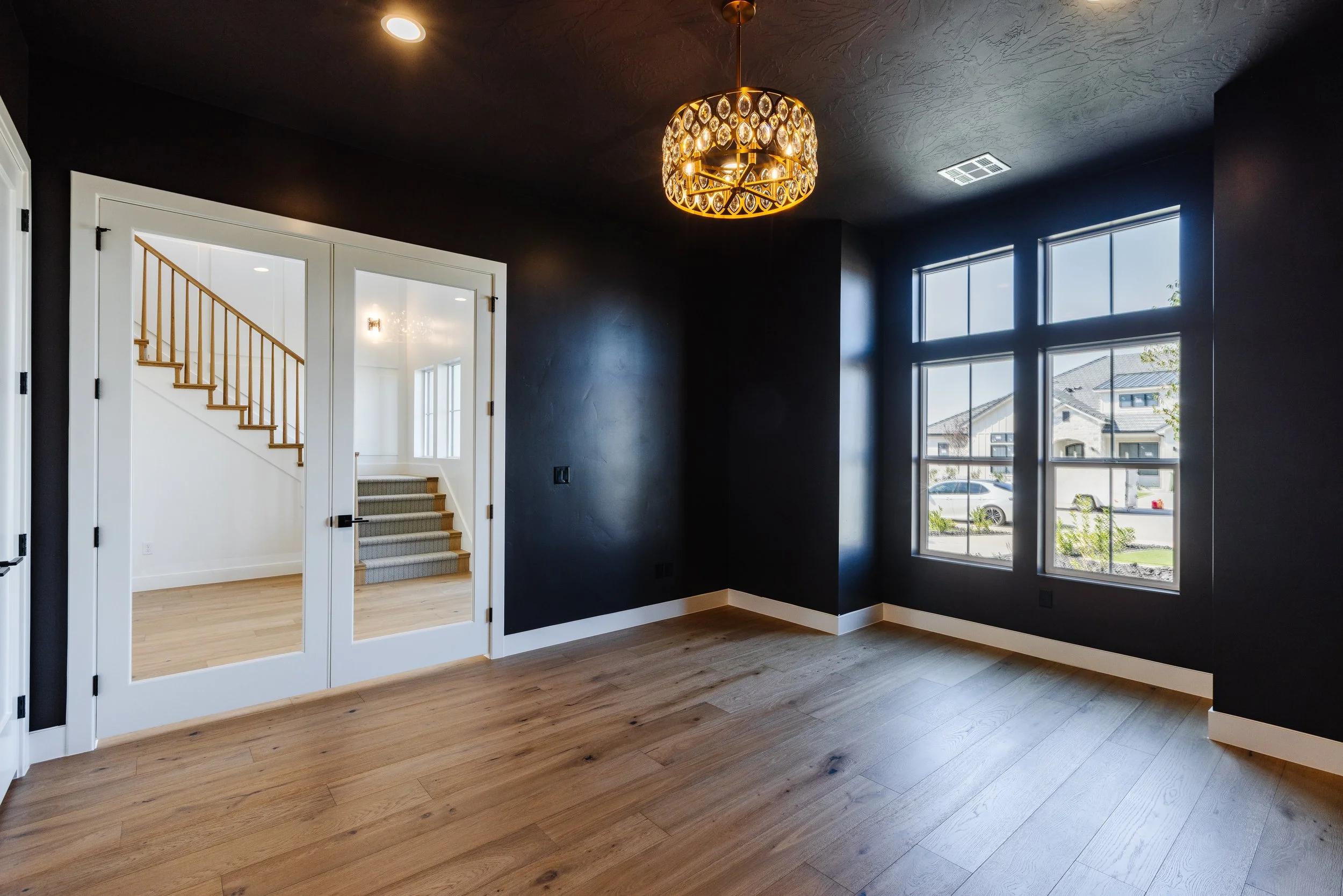 A modern living room with dark-colored walls, light hardwood flooring, and large windows letting in natural light. There is a decorative chandelier hanging from the ceiling, and a doorway with glass panels leads to a bright staircase area.