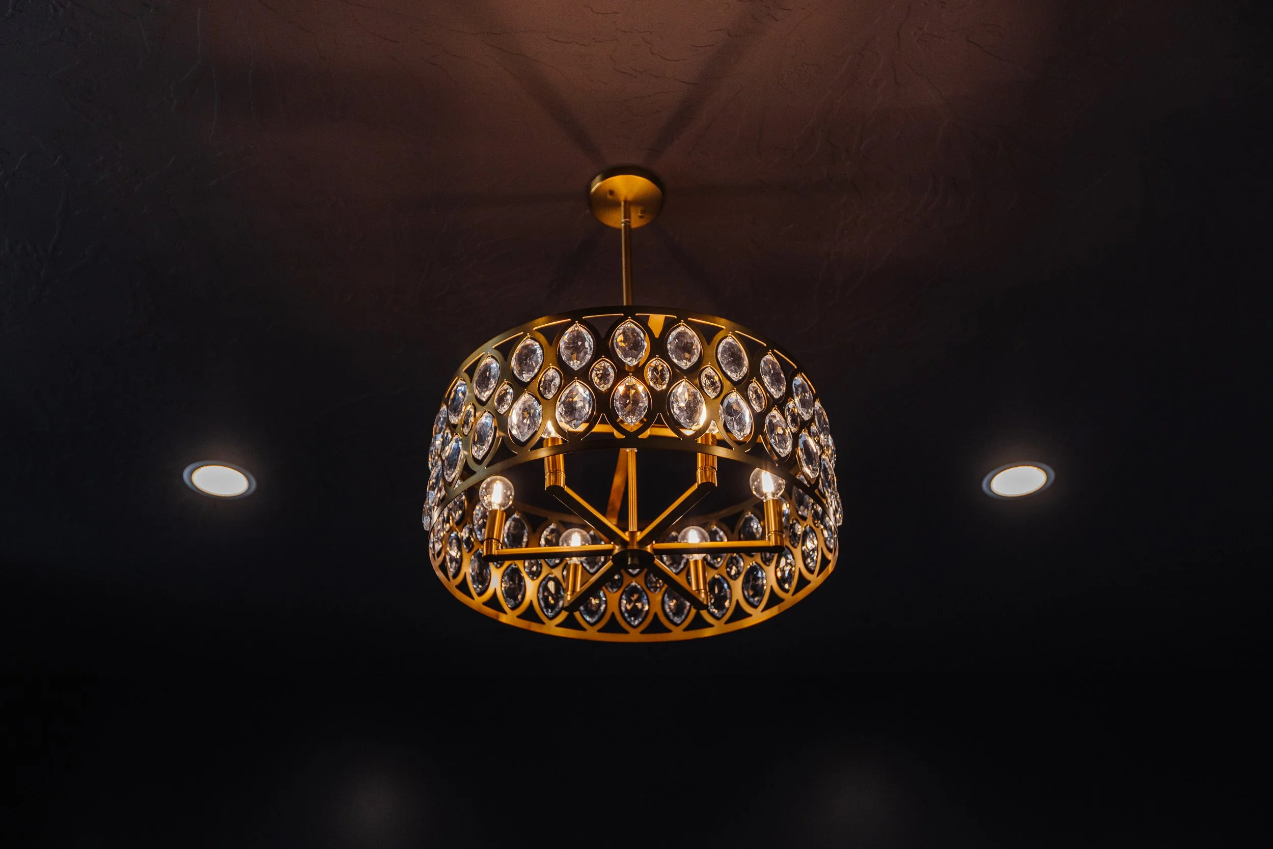 A decorative chandelier with crystal embellishments hangs from a dark ceiling, illuminated by nearby recessed lights.