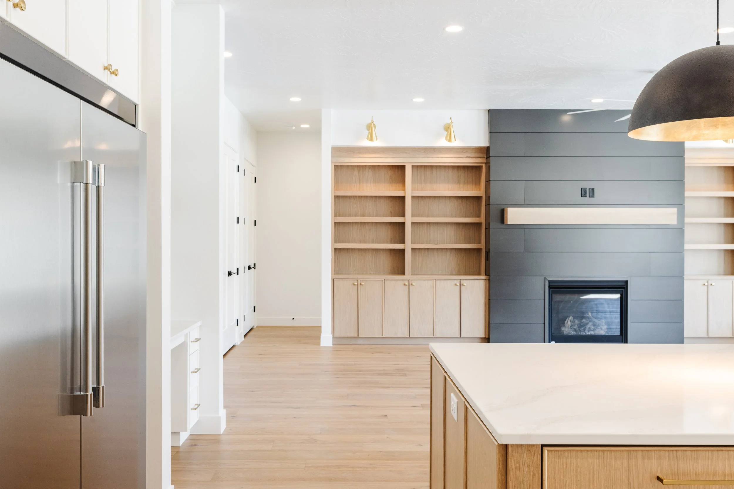 Modern kitchen with white walls, light wood flooring, built-in wooden shelving, gray shiplap wall with a fireplace, stainless steel refrigerator, and a kitchen island with a white countertop.