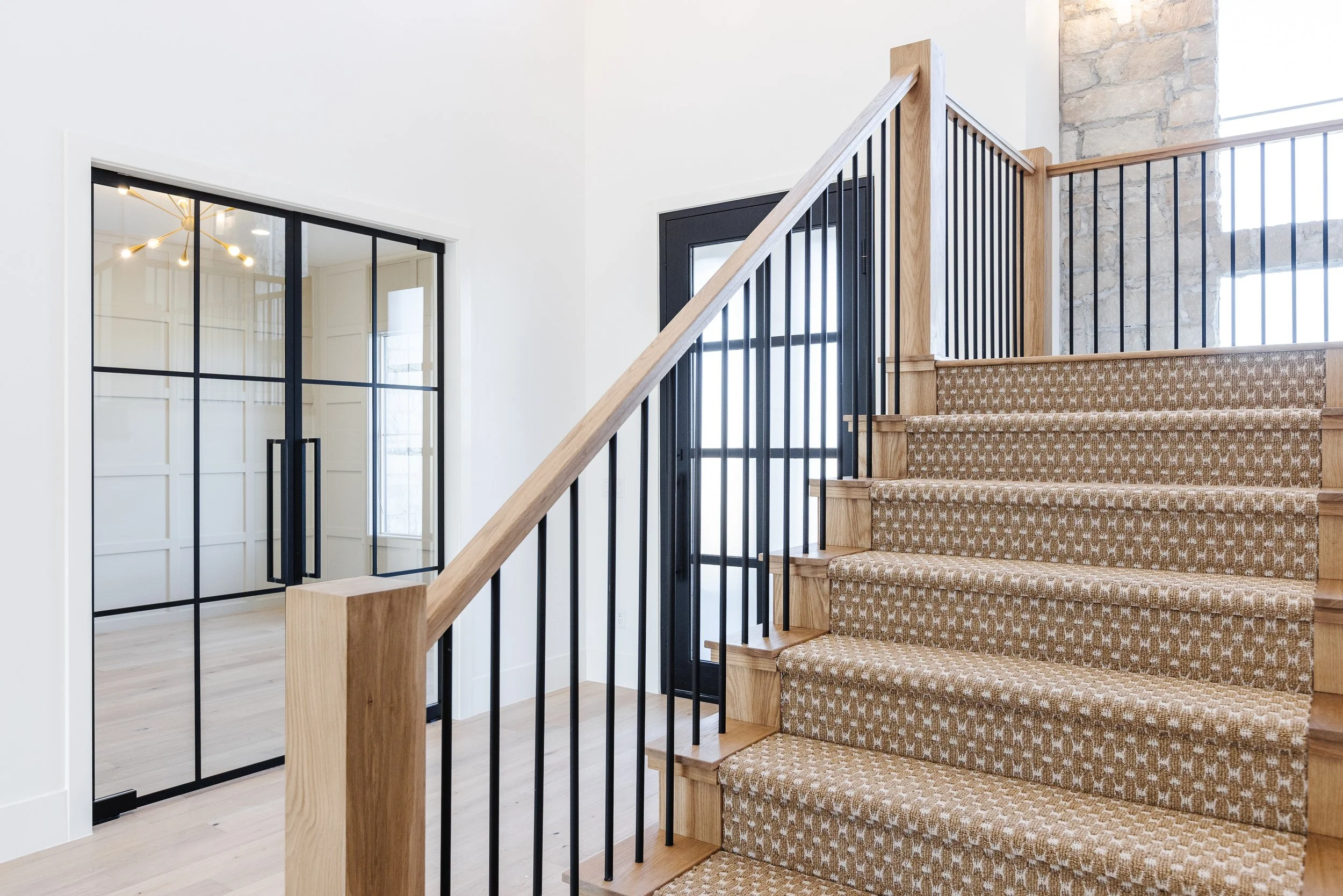 Interior of a modern home with a staircase featuring patterned carpeted steps, wooden handrail, and black metal balusters. Adjacent black-framed glass doors lead to a room with white walls and a gold chandelier, and exposed brickwork is visible near 
