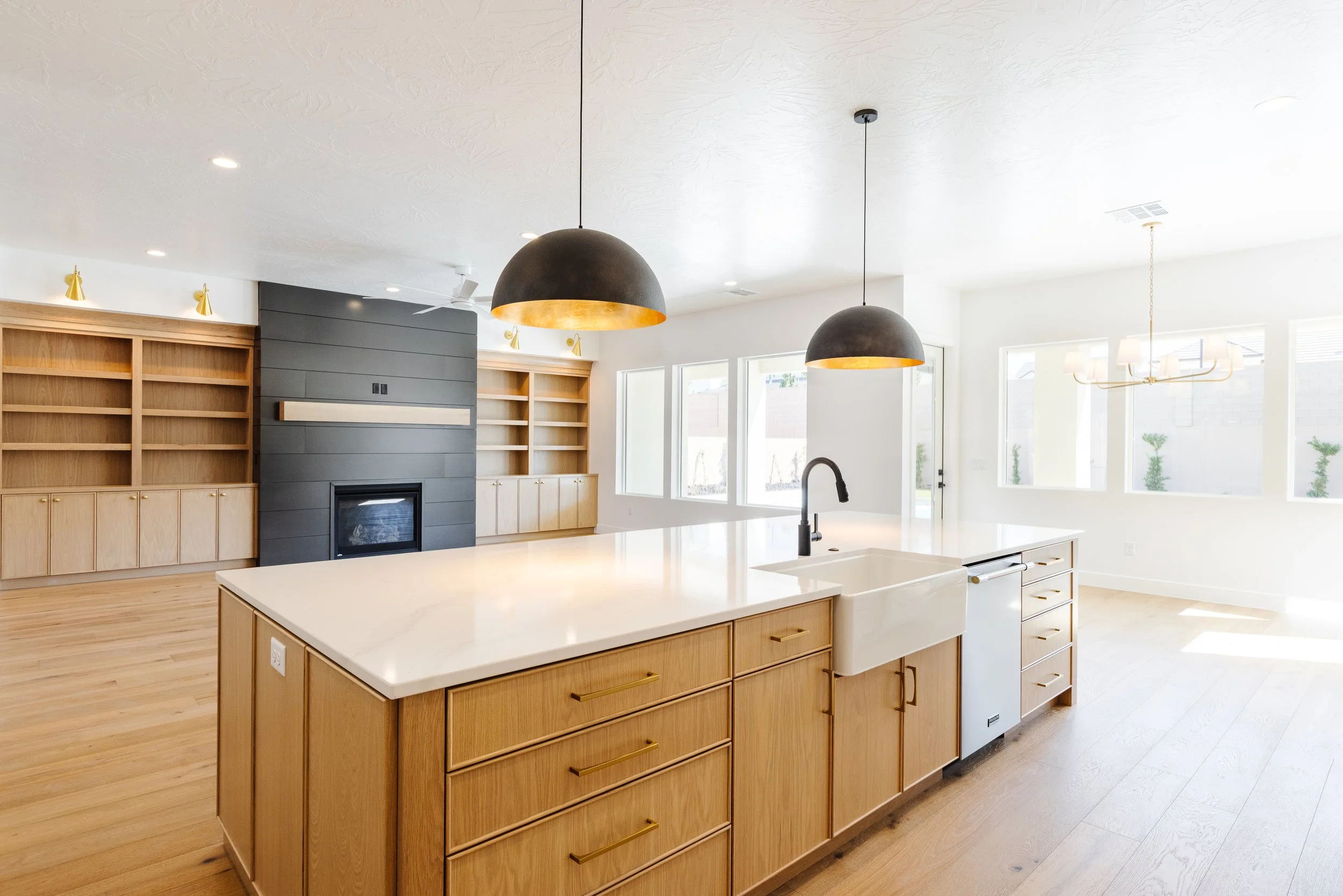 Modern kitchen with white island, black pendant lights, built-in wooden shelves, and large windows letting in natural light.