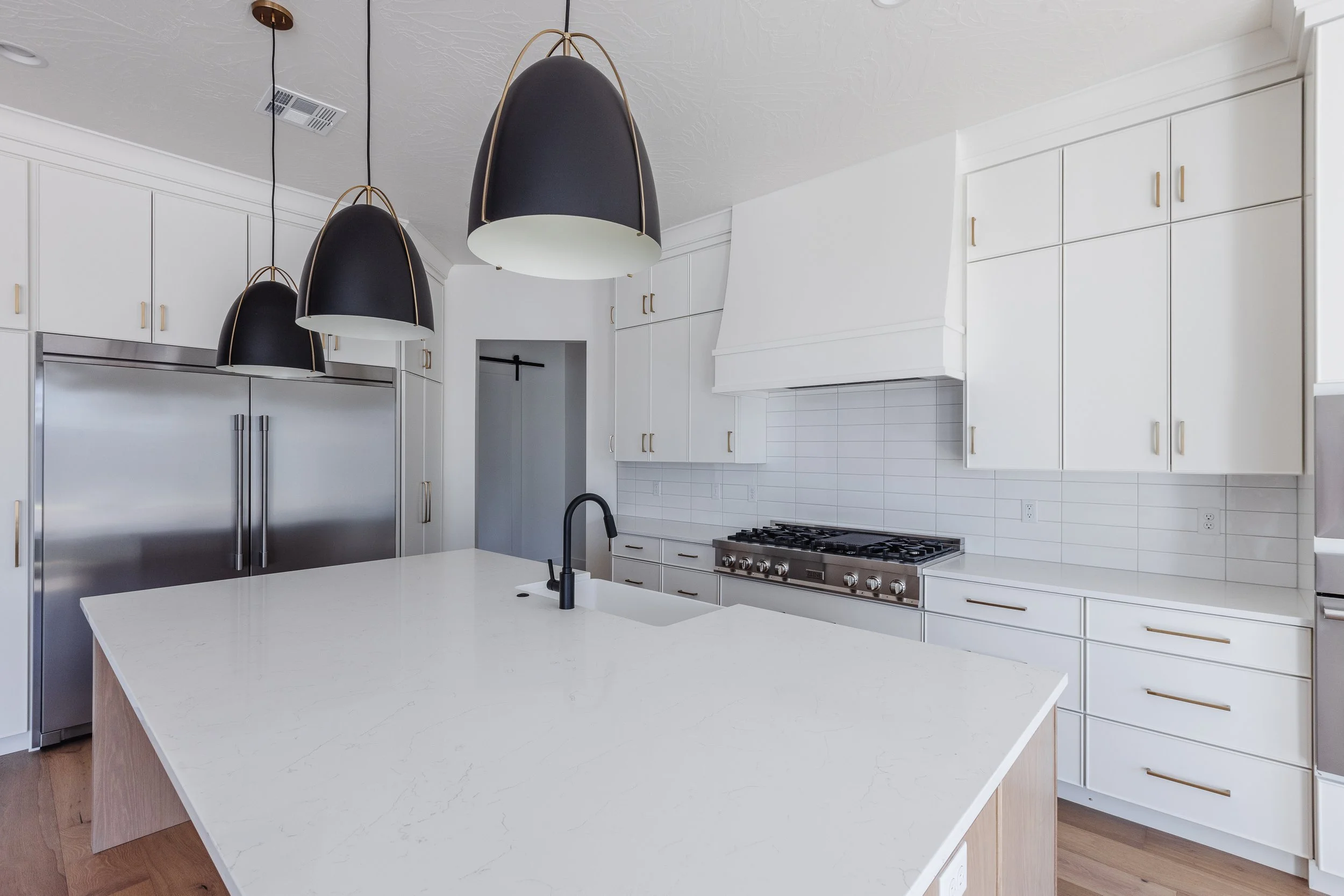 Modern white kitchen with black pendant lights, stainless steel refrigerator, gas stove, and an island with a white countertop.