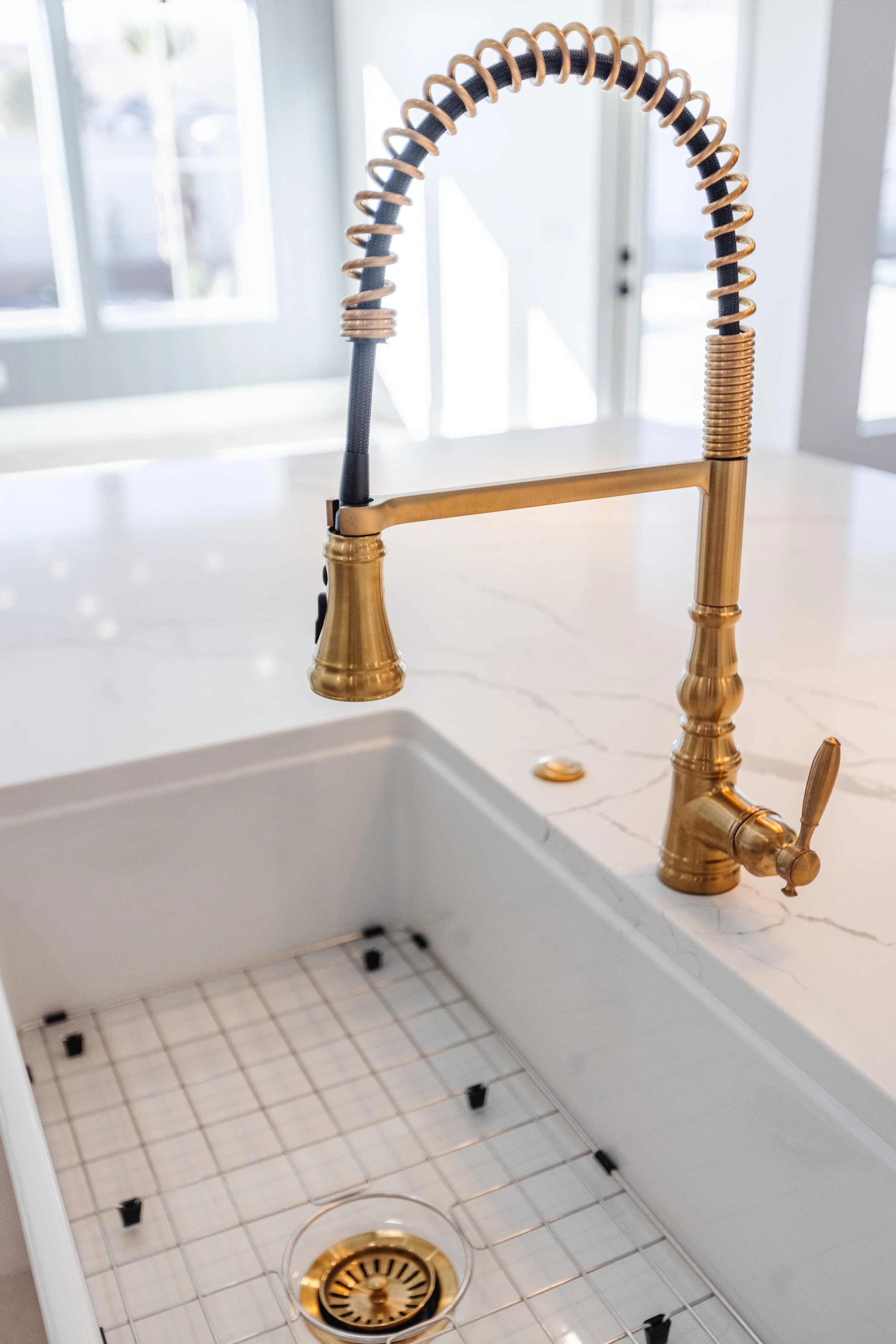 A kitchen sink with a gold-tone faucet, with the sink removed, exposing plumbing and drain. The countertop is white with a marble pattern, and a window with white curtains is in the background.