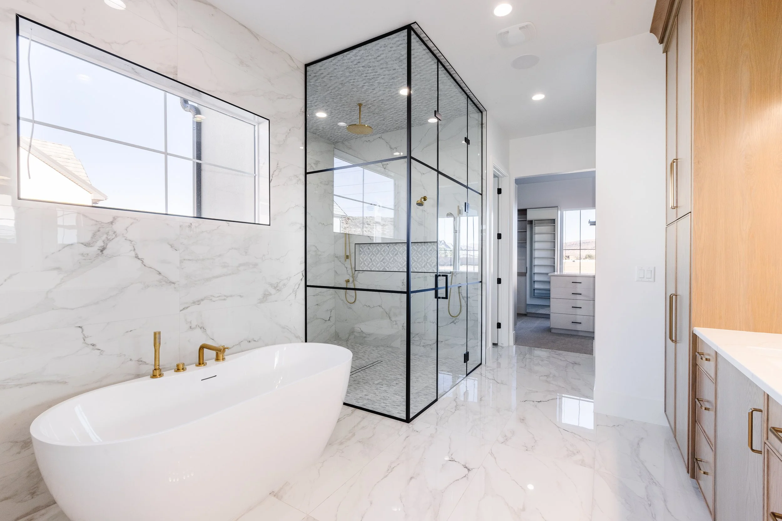 Modern bathroom with a freestanding bathtub, marble walls and flooring, a framed glass shower enclosure, and a wooden cabinet with gold handles.