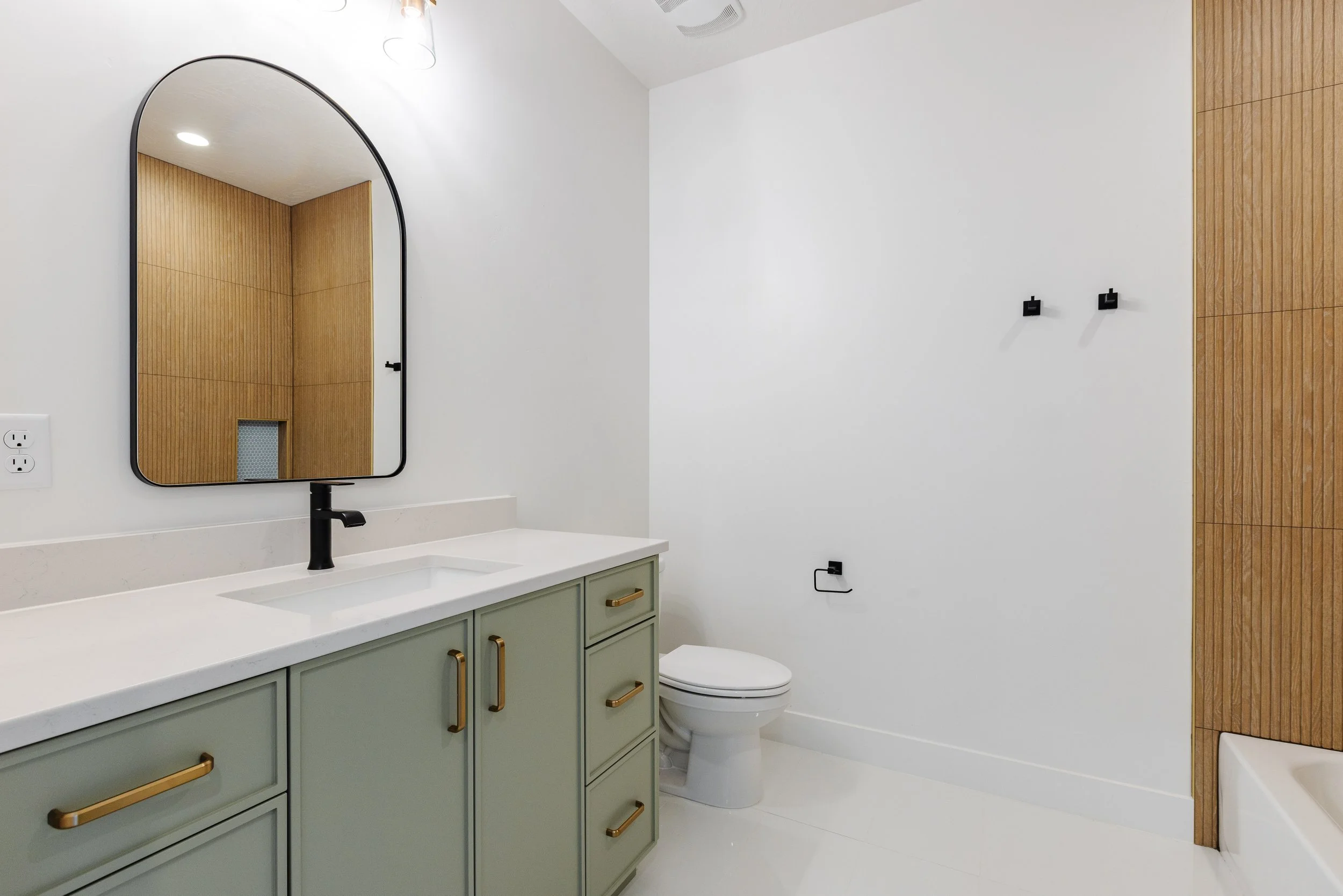 A modern bathroom with a light green vanity with gold handles, a white countertop and sink, a black faucet, a large arched mirror, a toilet, wooden wall paneling, and a white wall with black hooks.
