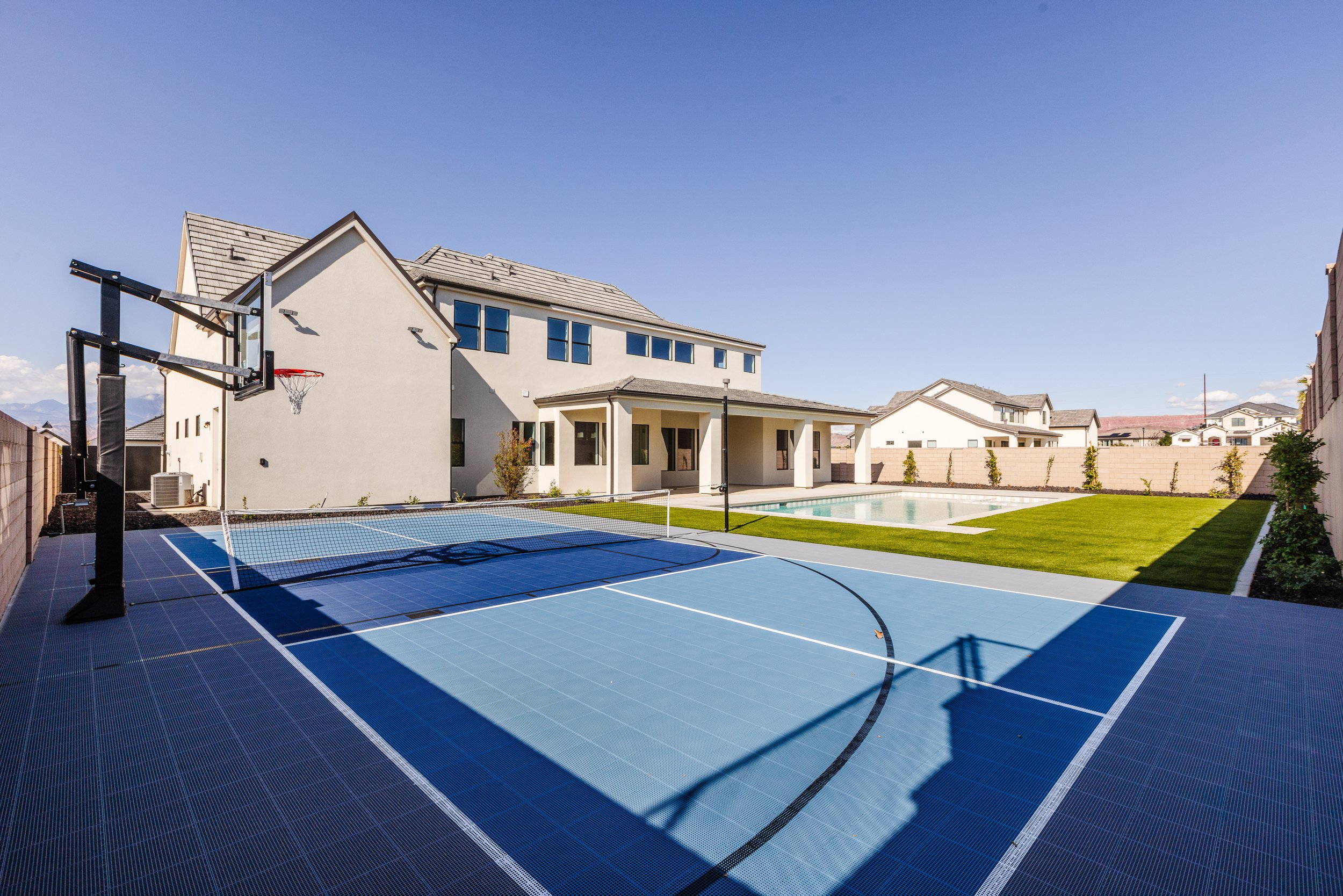 Backyard with a basketball hoop, a swimming pool, and a tennis court, with modern house and fence under a clear blue sky.