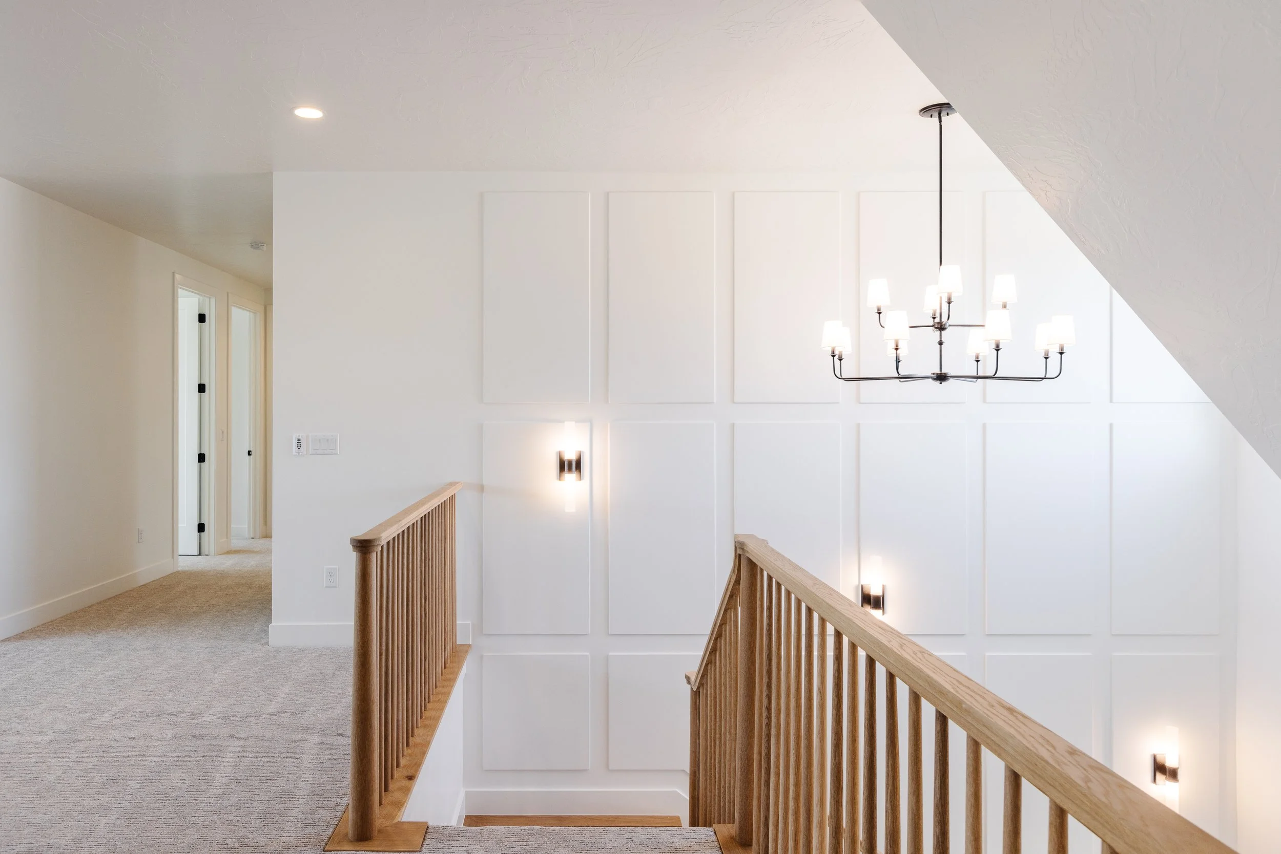 Interior view of a staircase landing with a hanging chandelier, white paneled wall, wall-mounted light fixtures, and white doors leading to different rooms.